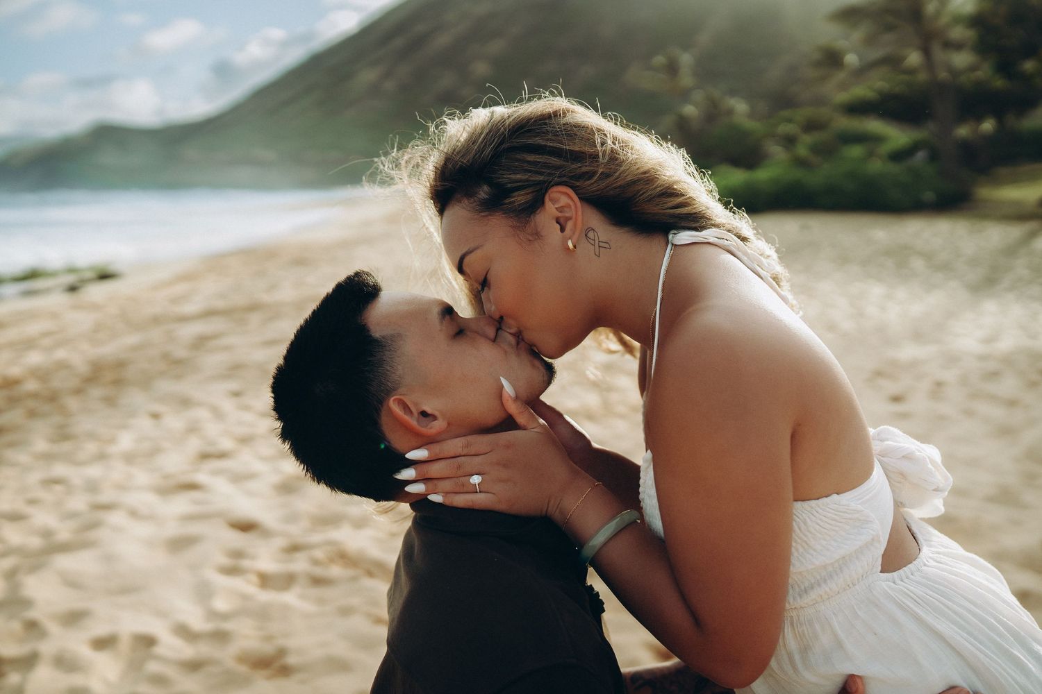 Couple sharing a romantic kiss on a sandy beach with lush green mountains and golden sunlight in the background at Sandy beach Oahu Hawaii