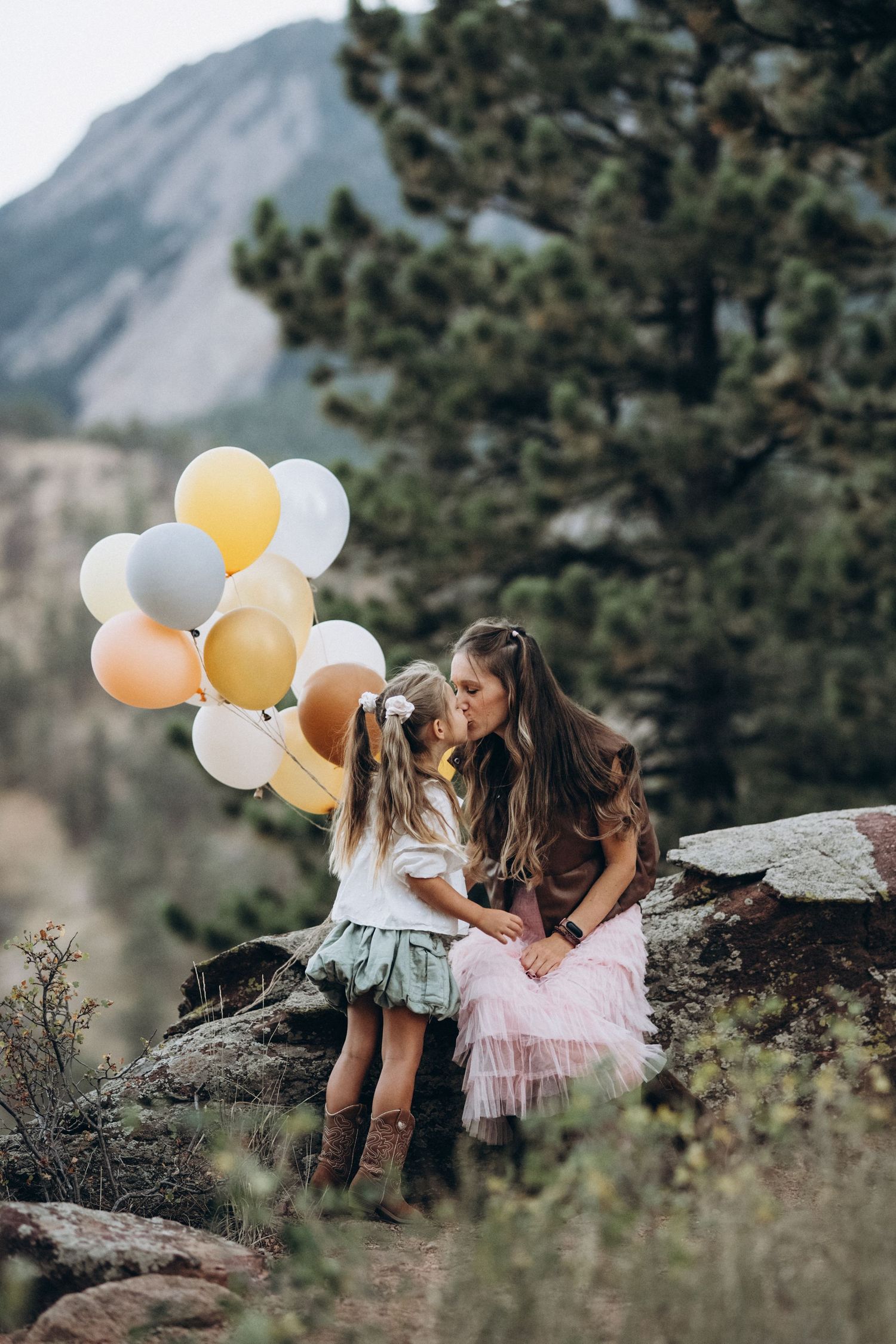 Mom and daughter sitting together with balloons by a pine tree and rocks, Boulder family photographer at NCAR, Colorado