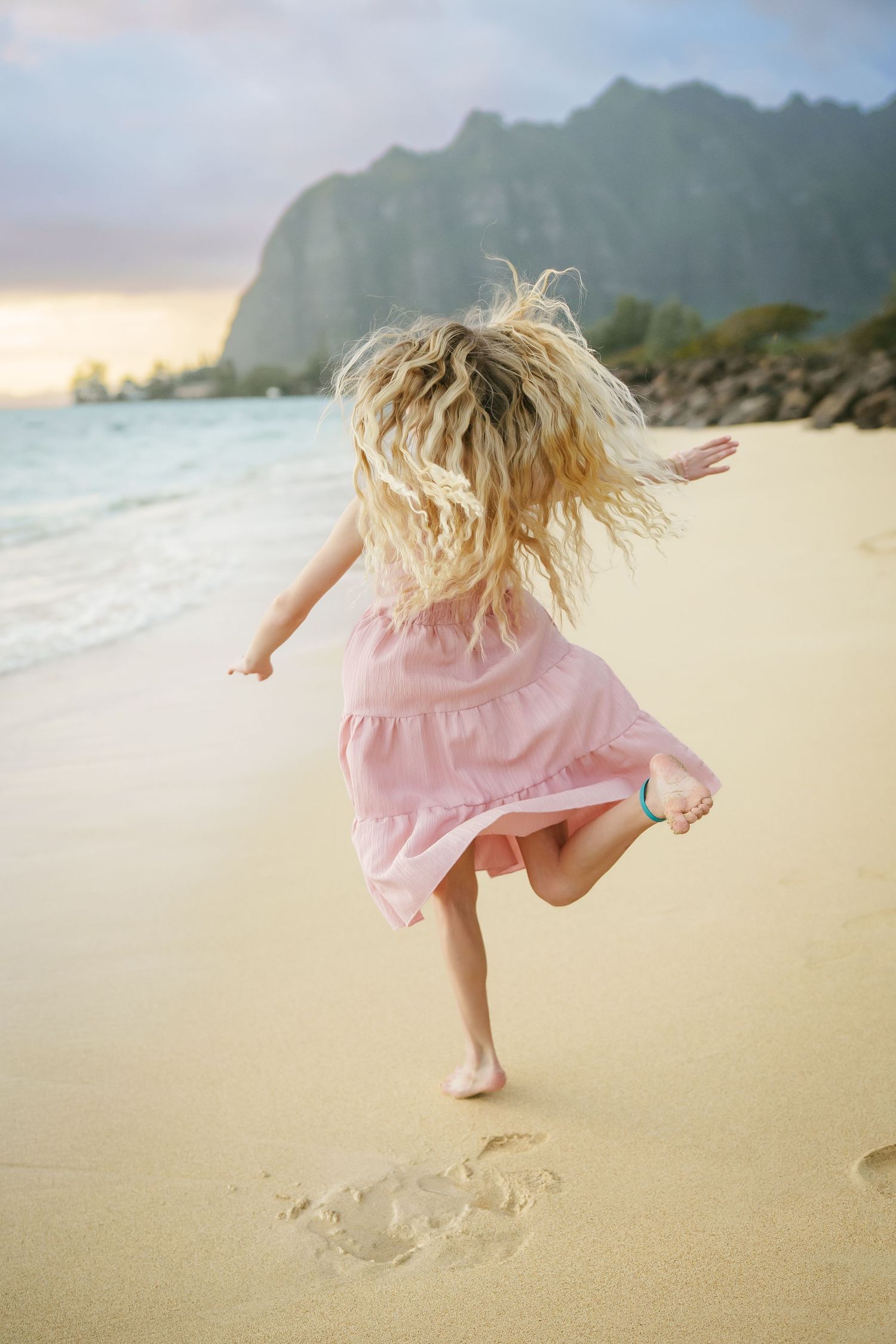 A girl in a pink dress running along the beach at sunrise with mountains in the background. Cinematic shot captured at Ka’a’awa Beach, Oahu.