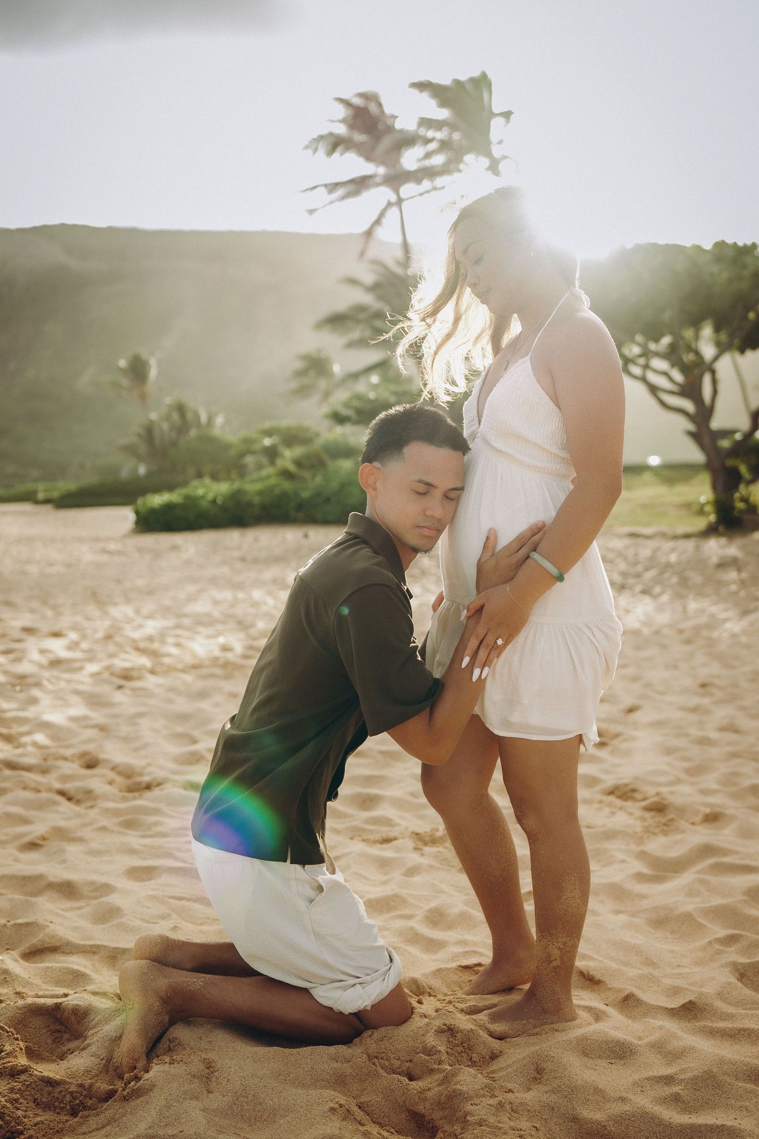 Man kneeling on sandy beach pressing ear to pregnant woman's belly during golden sunset in Hawaii at Sandy beach Oahu Hawaii