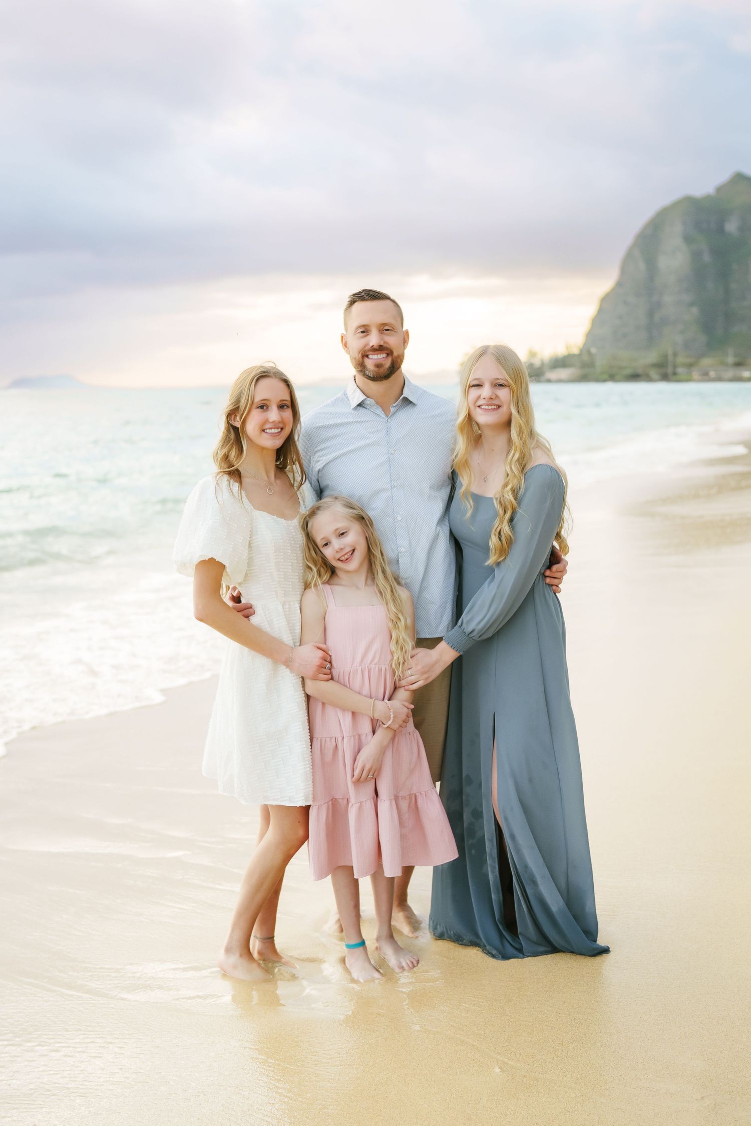 A tender full-length portrait of a father and three daughters hugging and smiling at sunrise on Ka’a’awa Beach, Oahu. Scenic mountains behin