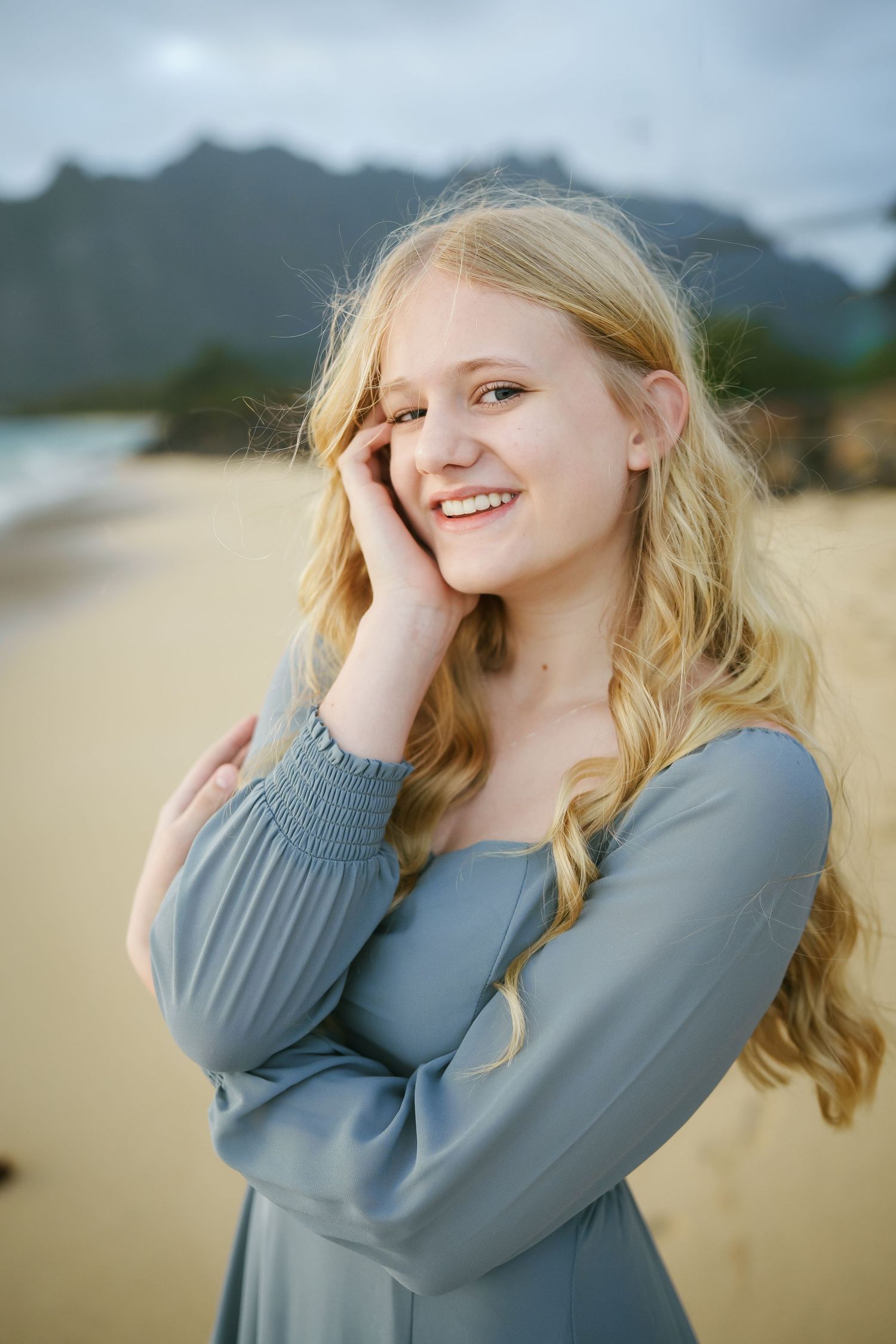 A beautiful blonde girl smiling and posing naturally at Ka’a’awa Beach, Oahu. Scenic mountains in the background during a photoshoot.