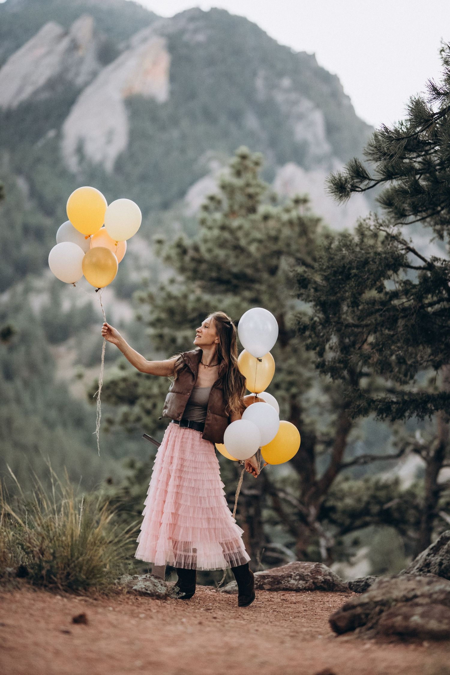 Girl in pink tulle skirt reaching for yellow balloons among pine trees, Boulder family photographer, NCAR, Colorado