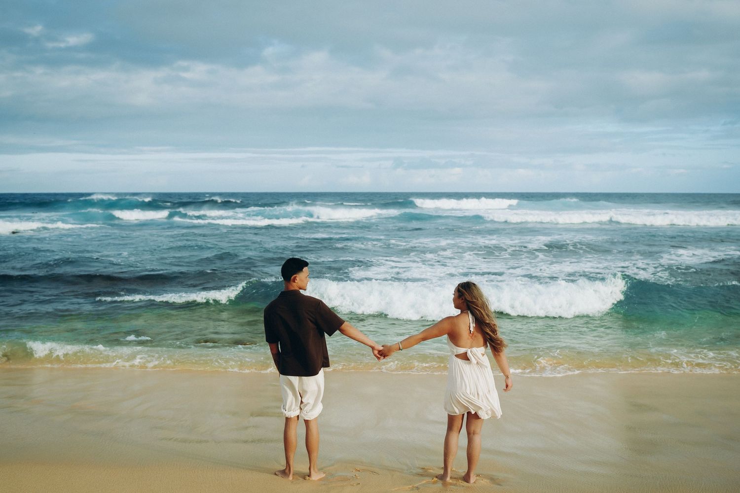 Couple holding hands on a sandy beach facing turquoise ocean waves under a cloudy sky at Sandy beach Oahu Hawaii