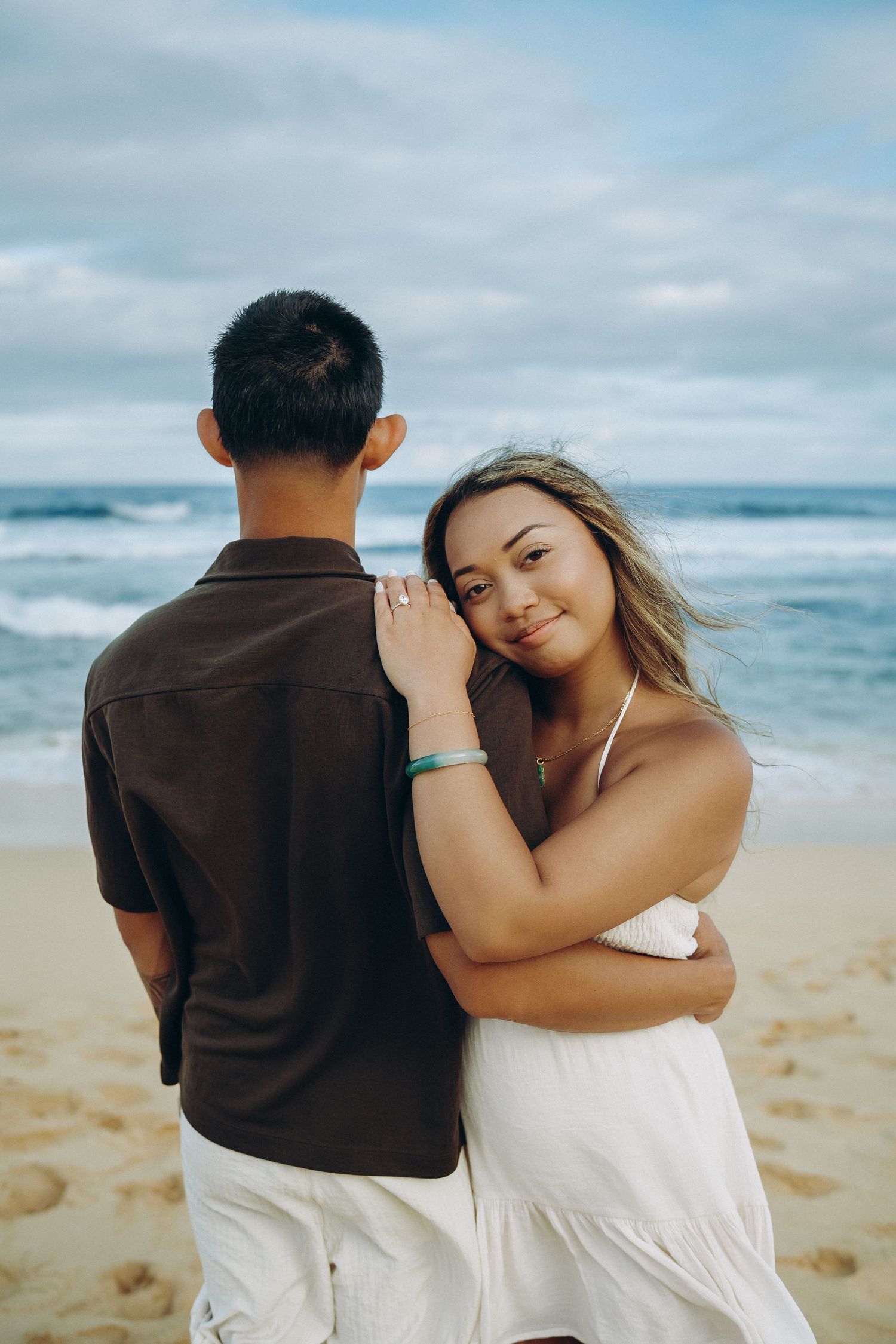A couple embraces on a sandy beach, the woman in a white dress smiling over her partner's shoulder with ocean waves behind them. Anna Tsenta