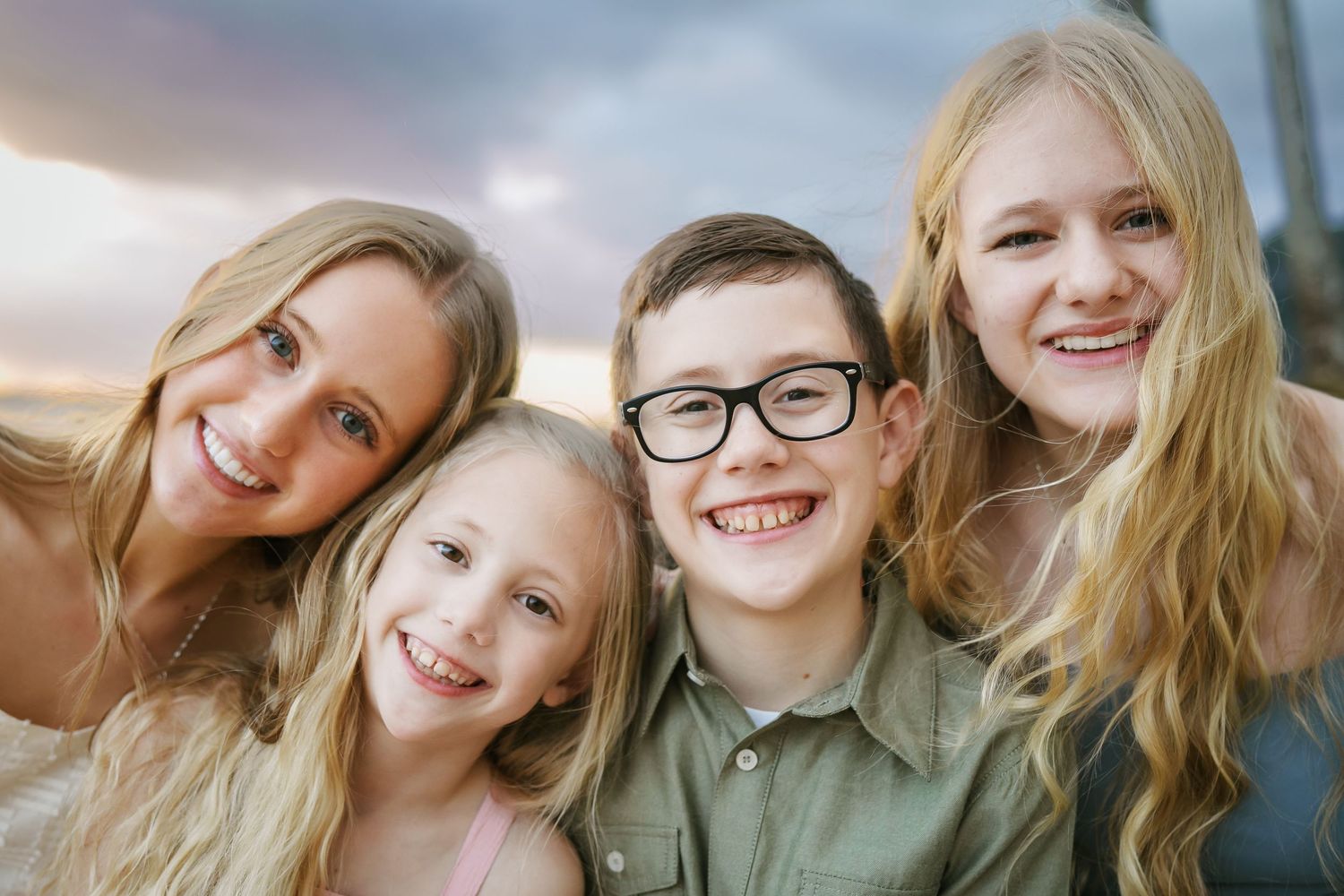 Close-up portrait of four siblings: three blonde sisters and their brother in glasses. All smiling happily against a beautiful Oahu sunrise.