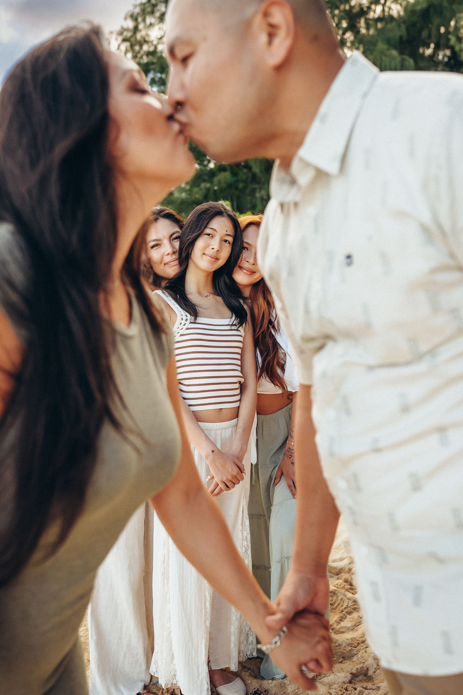 Secret Beach family photoshoot, Oahu. Parents sharing a kiss while kids stand behind them. Captured at scenic Kualoa Ranch area.