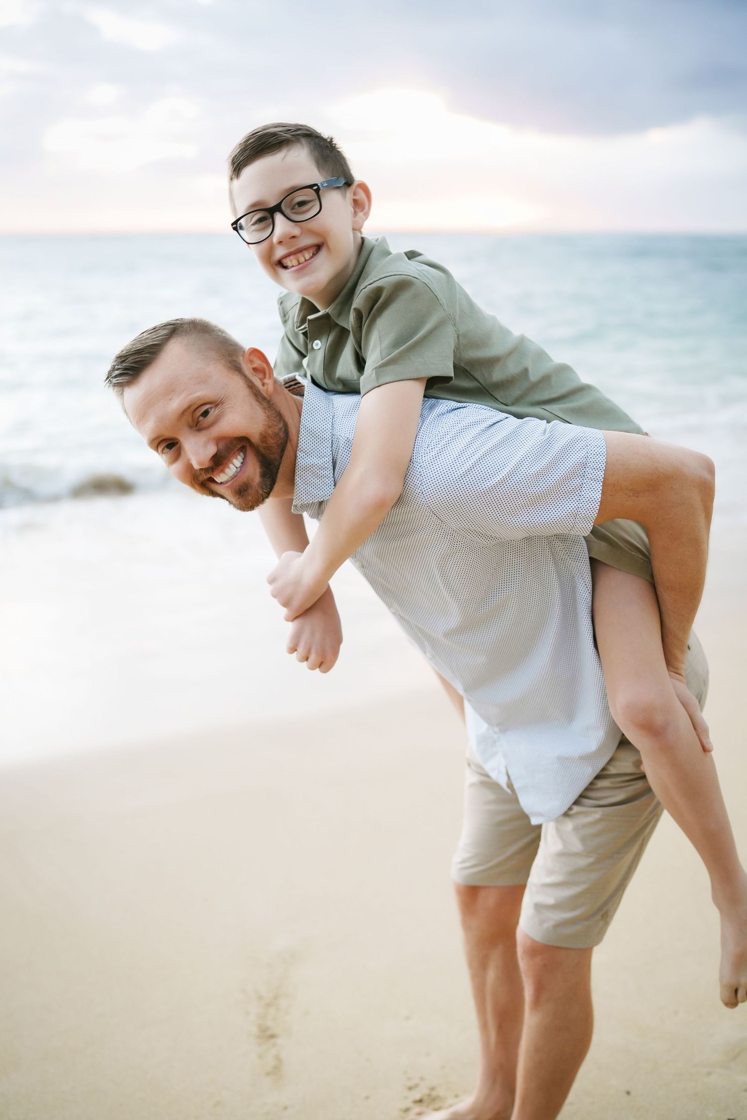 A playful moment with a son riding on his father's back at sunrise. They are laughing and fooling around with the ocean in the background
