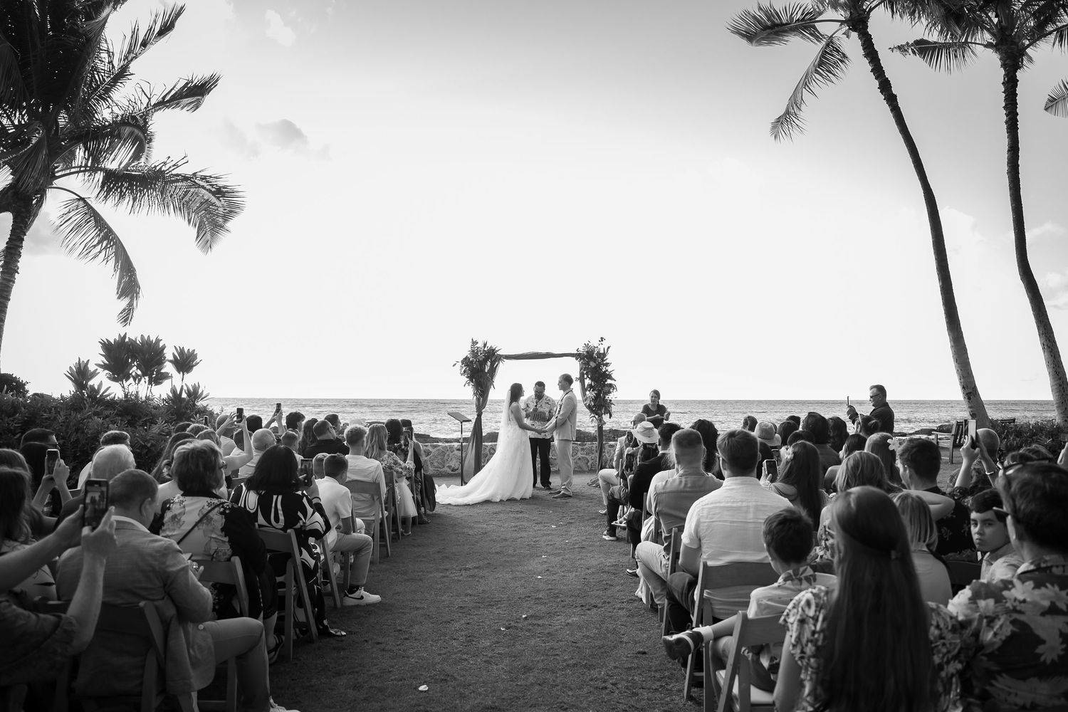 B&W wide shot of a beach wedding ceremony in Ko Olina. Guests seated facing the ocean. Timeless Oahu destination wedding photography.