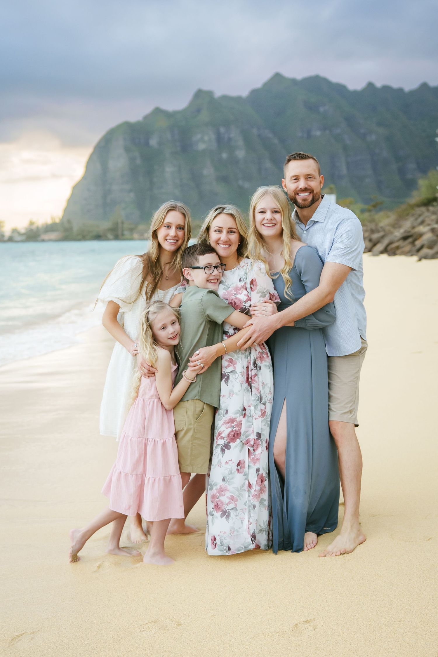 A family poses together on a beautiful Kaaawa beach with dramatic mountains of Kualoa in the background at sunset.