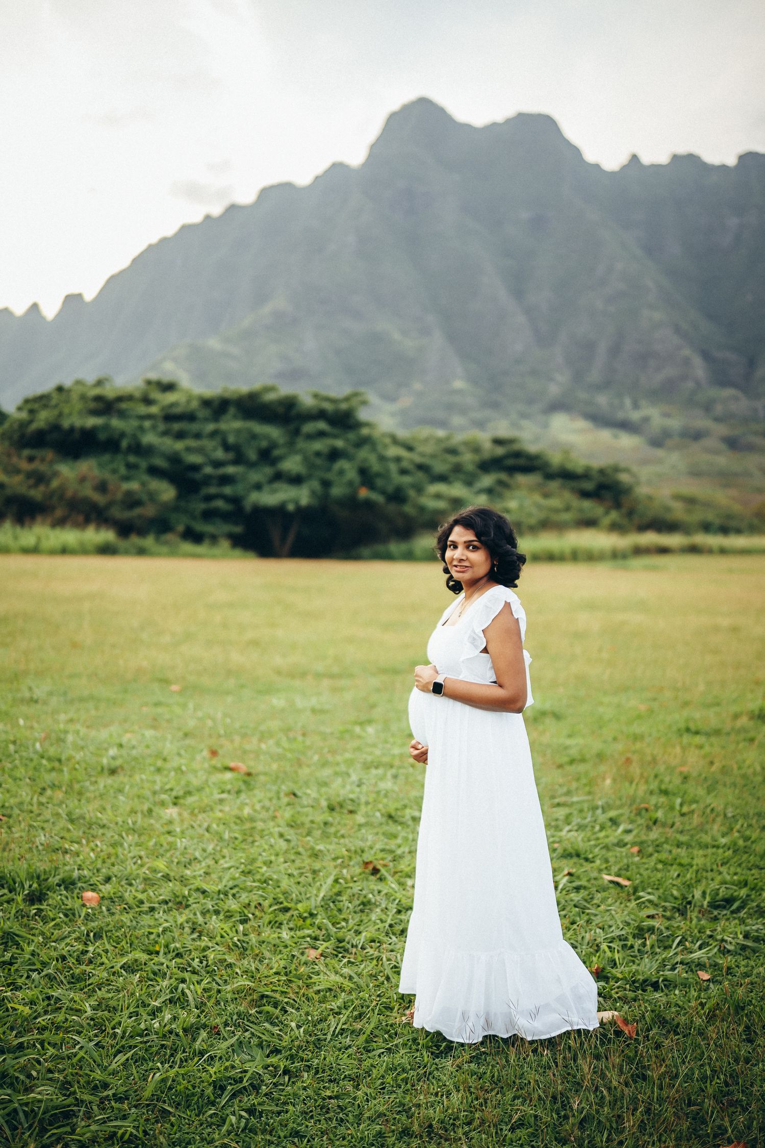 Maternity family photoshoot, Oahu. A pregnant woman in a white dress standing in a field at Kualoa Ranch near Secret Beach. Nature vibes.