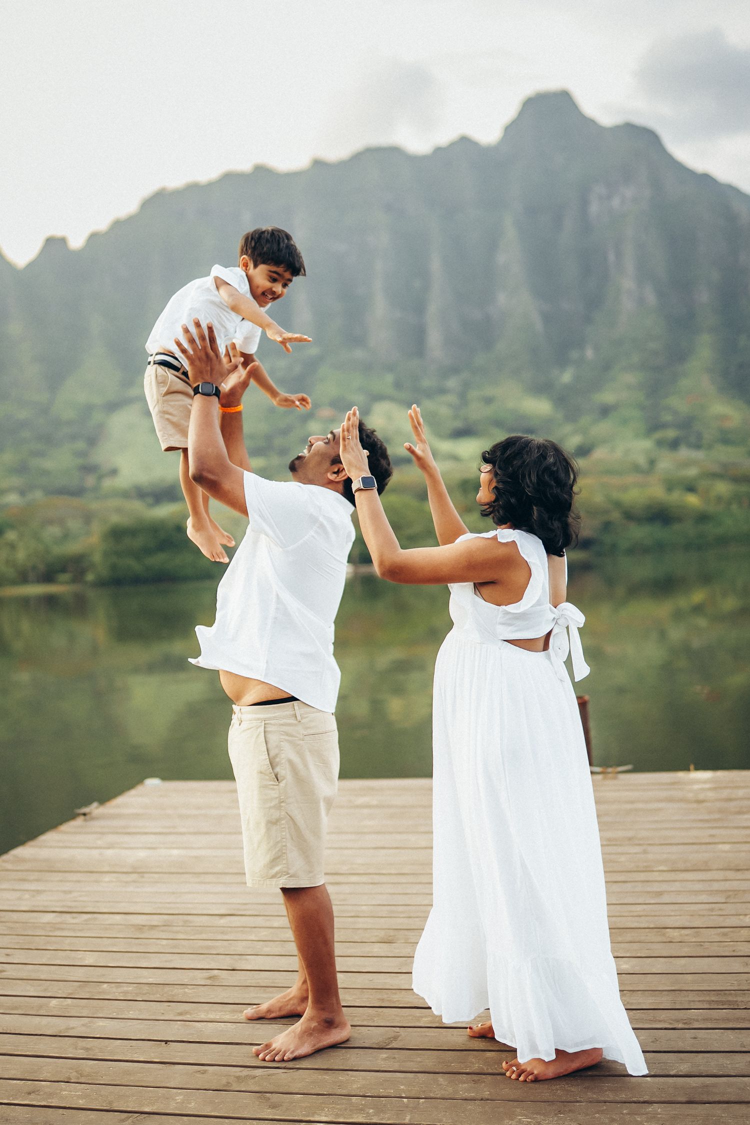 Joyful family photoshoot at Secret Beach. Parents and child on a dock near Kualoa Ranch, Oahu. Stunning mountain view photography.