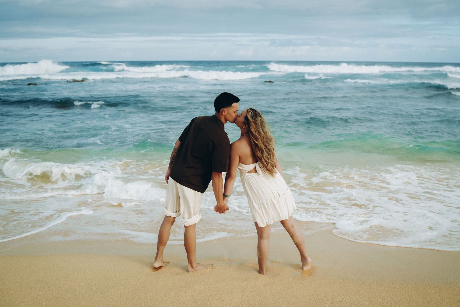 Couple kissing on a sandy beach while holding hands with ocean waves in the background at Sandy beach Oahu Hawaii