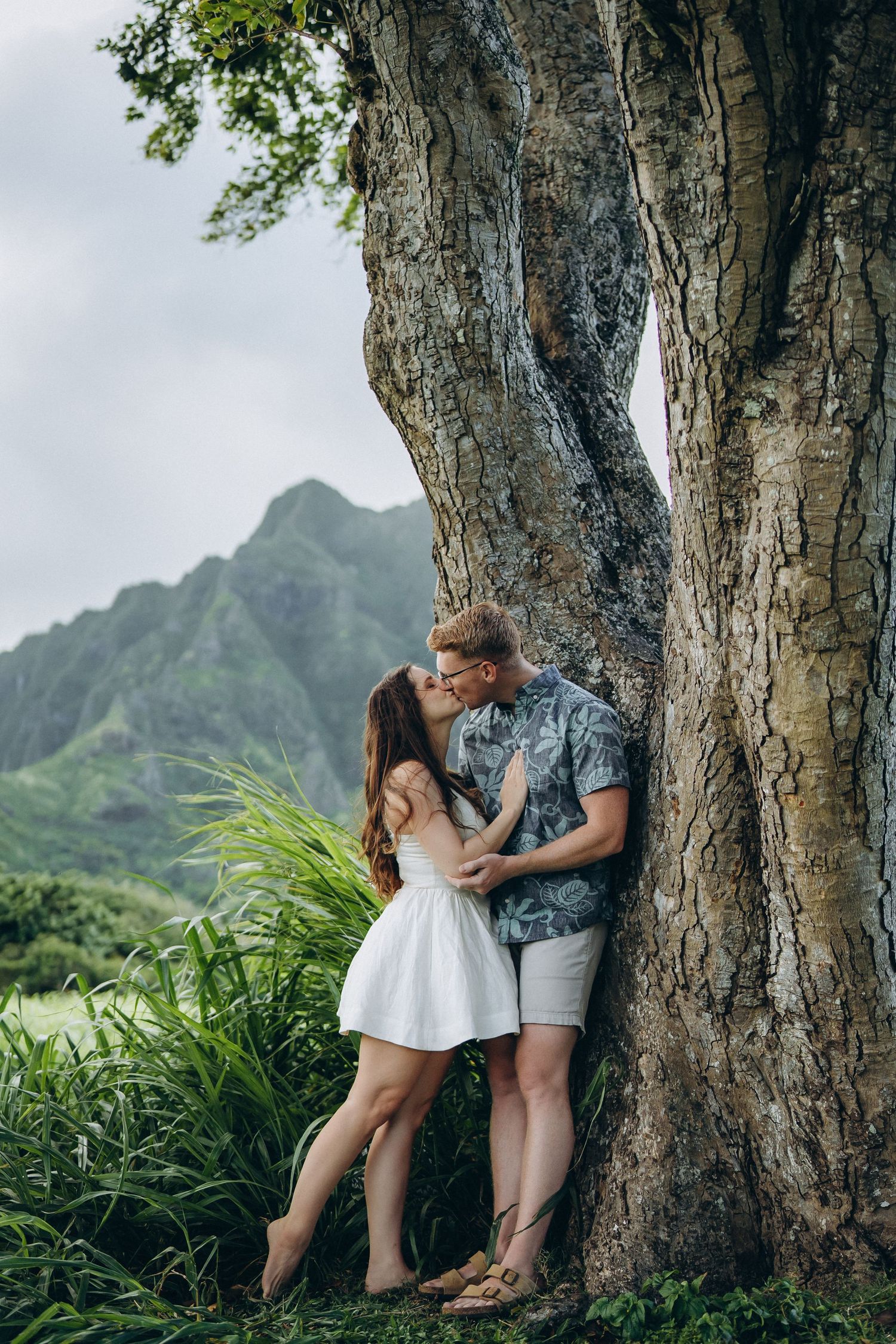 Couple embracing against a large tree trunk with mountains behind, Kualoa Ranch couples photographer, Oahu, Hawaii