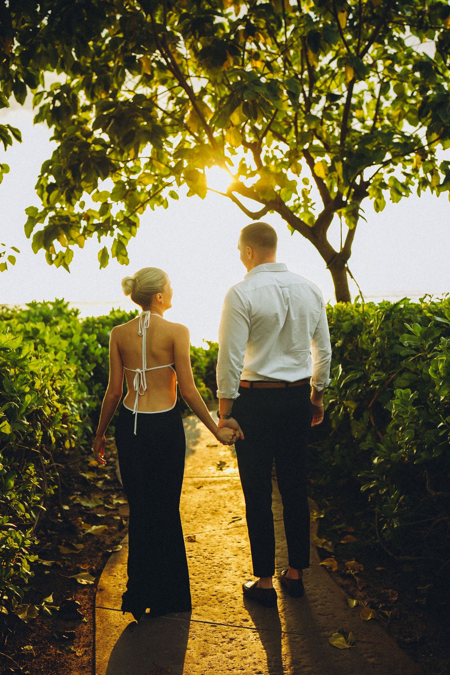 A couple walks hand in hand along a sunlit garden in Ko Olina path beneath a leafy tree during golden hour.