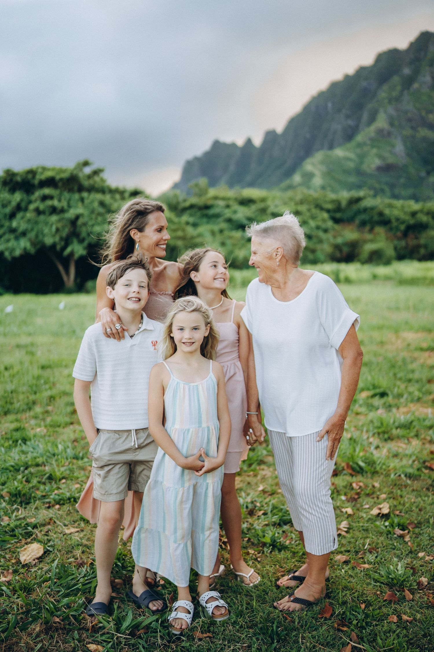 Multi-generational family of five smiling together on green lawn with dramatic mountain backdrop in Hawaii.