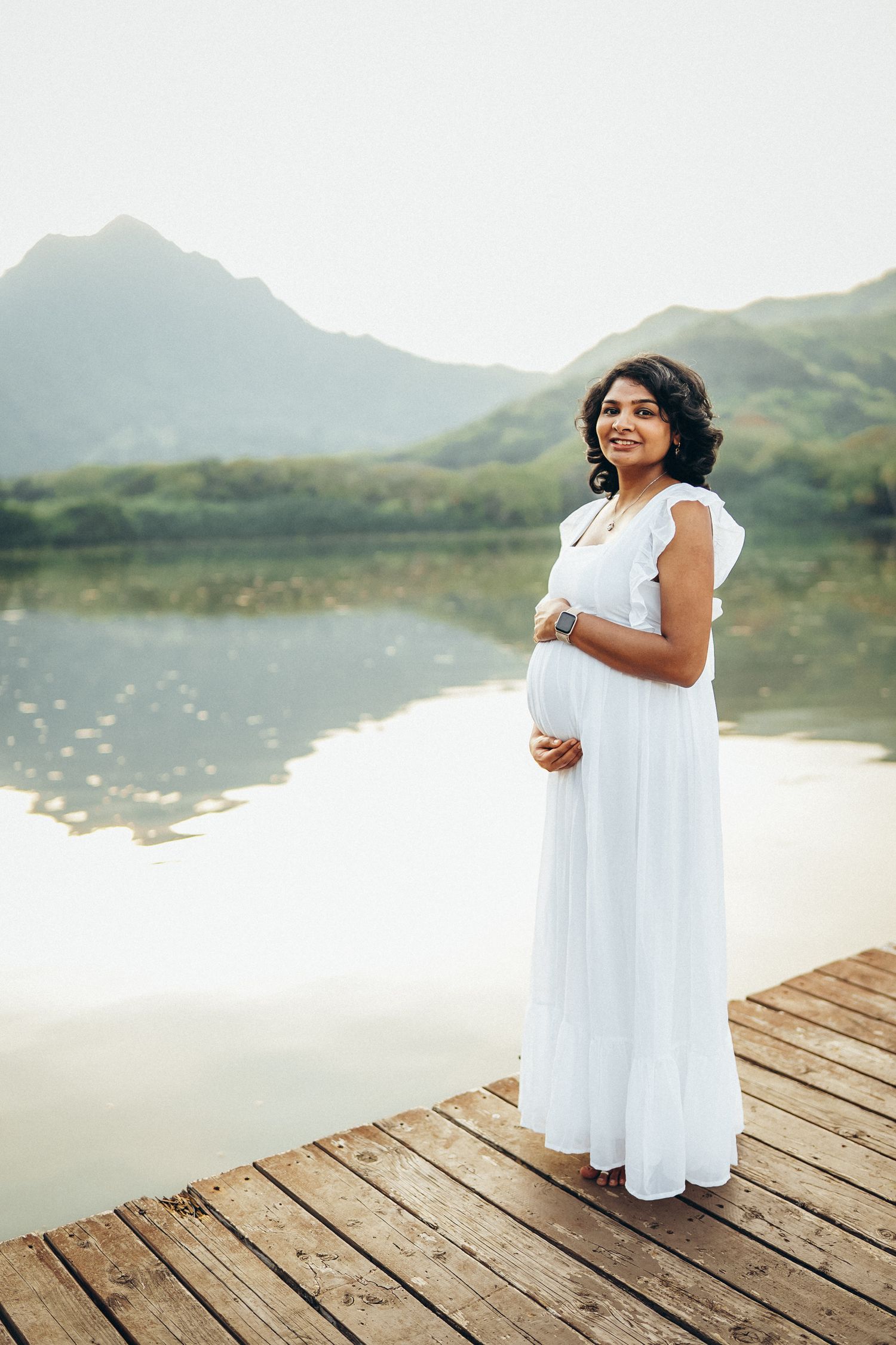 Beautiful Oahu family photoshoot. A pregnant woman in a white dress posing on a dock at Secret Beach near Kualoa Ranch.