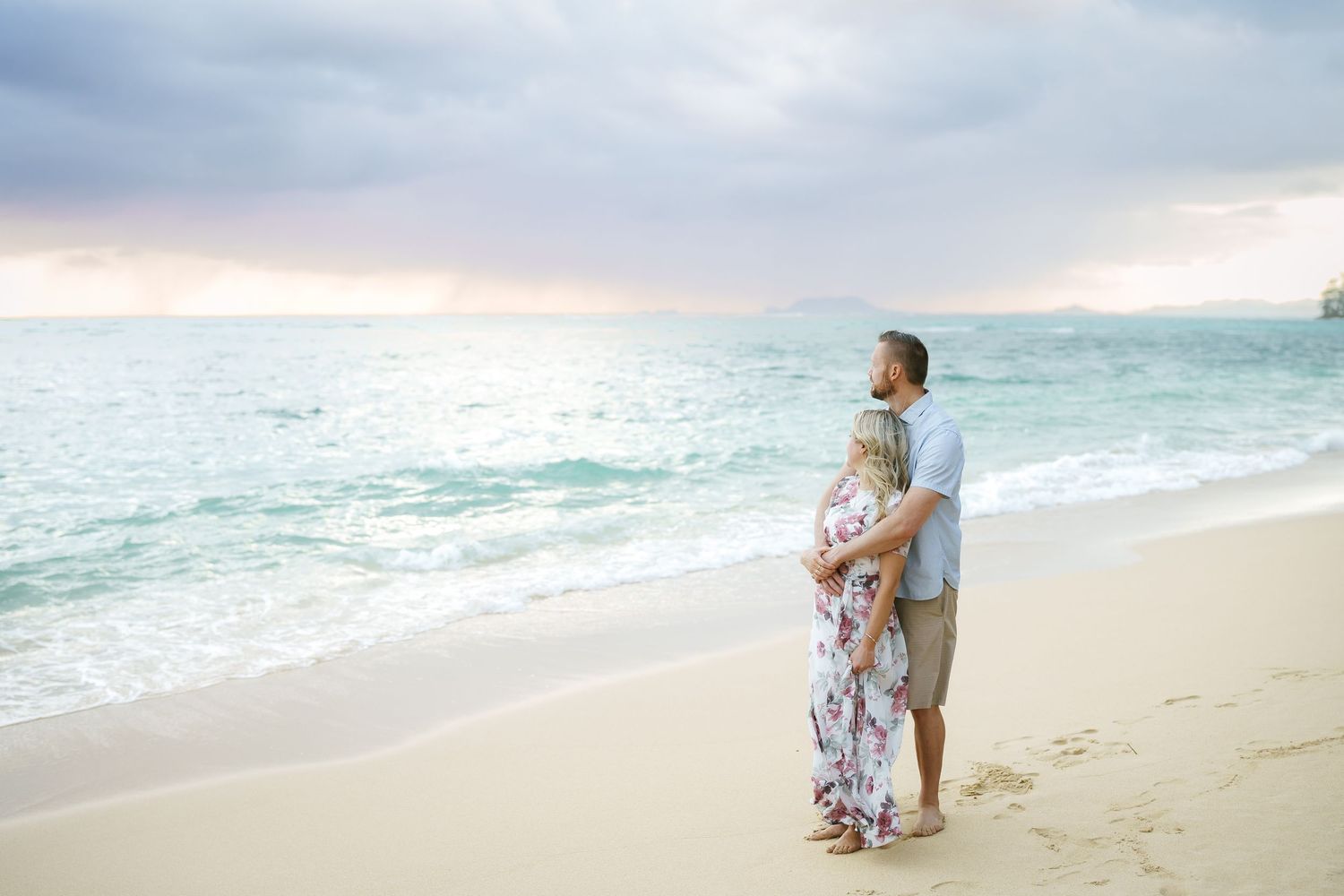 Parents embracing and watching the sunrise at Ka’a’awa Beach, Oahu. A romantic moment by the ocean during a family photoshoot in Hawaii.