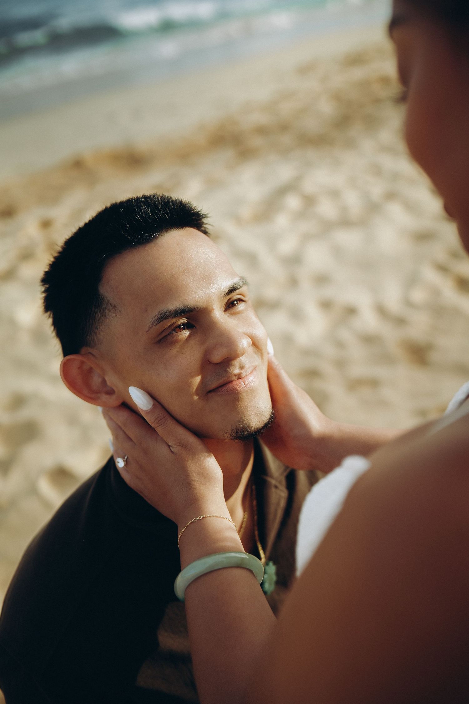 A pregnant woman applies sunscreen to a smiling man's face on a sandy beach with ocean waves in the background at Sandy beach Oahu Hawaii