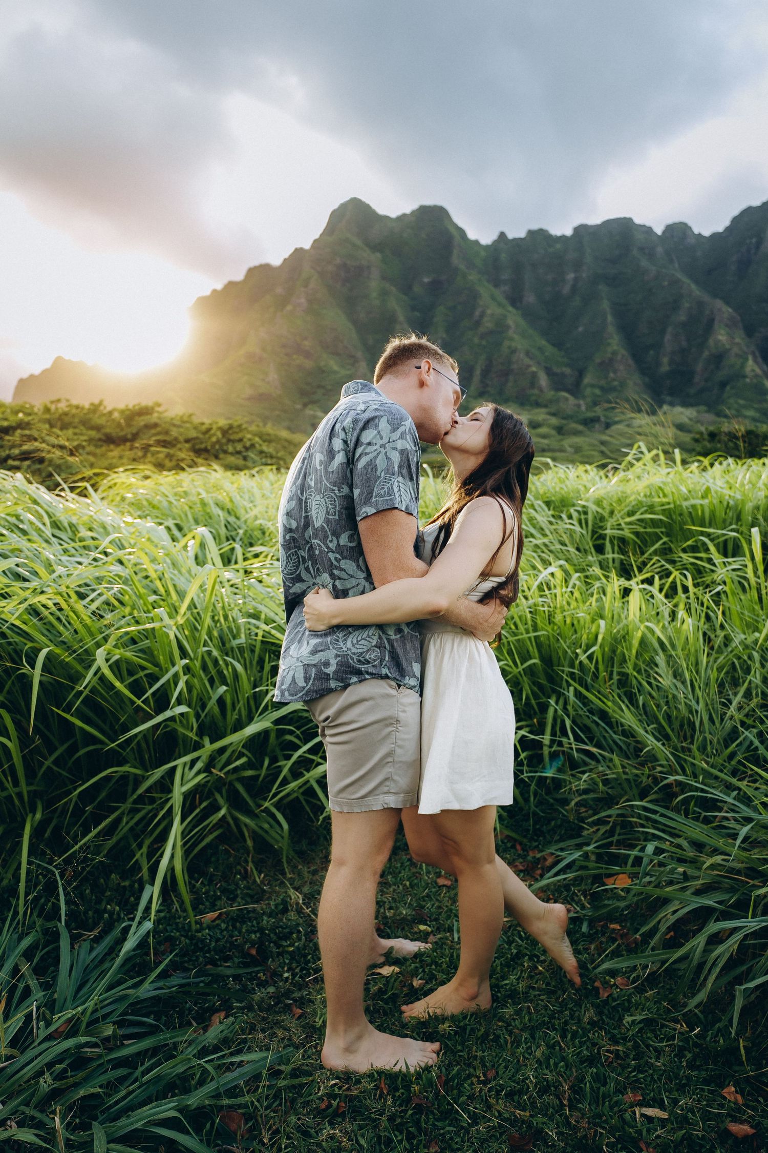 Couple kissing in tall green grass at golden hour, mountains behind, Kualoa Ranch Oahu couples photographer