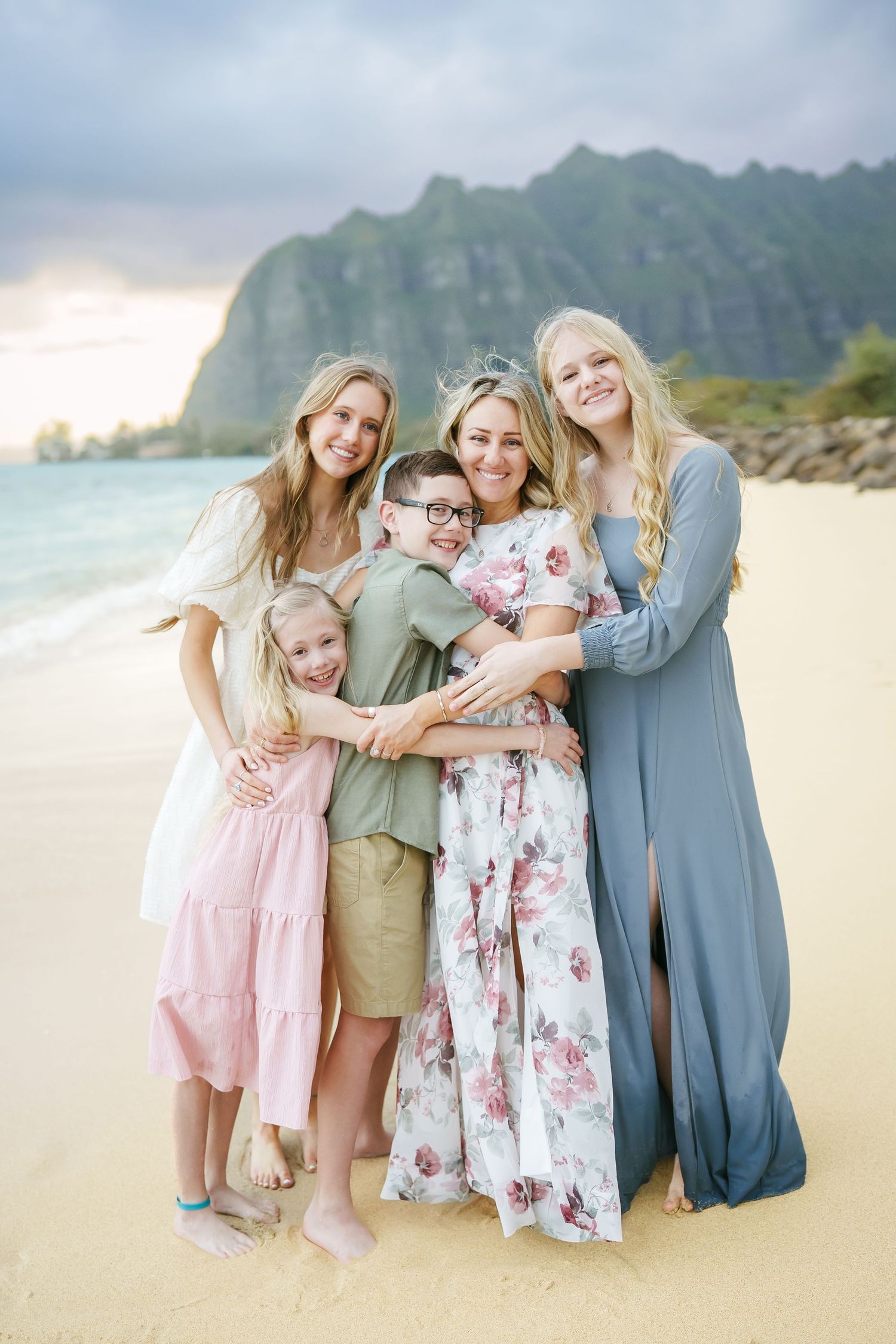 Four children smiling and hugging their mother at Ka’a’awa Beach, Oahu. A beautiful family moment on white sand with ocean waves behind.