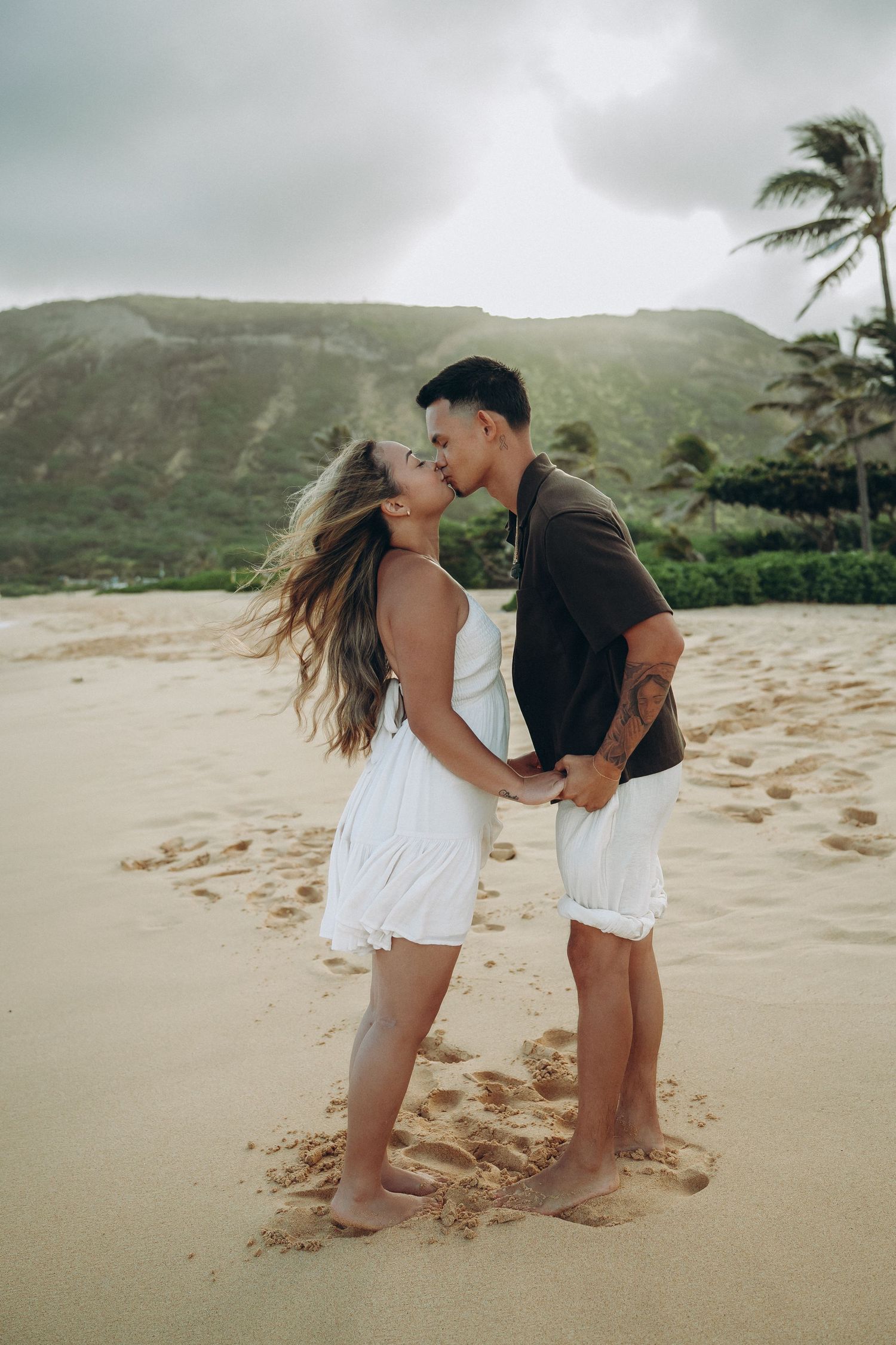 A couple kisses on a sandy beach with palm trees and green mountains in the background at Sandy beach Oahu Hawaii