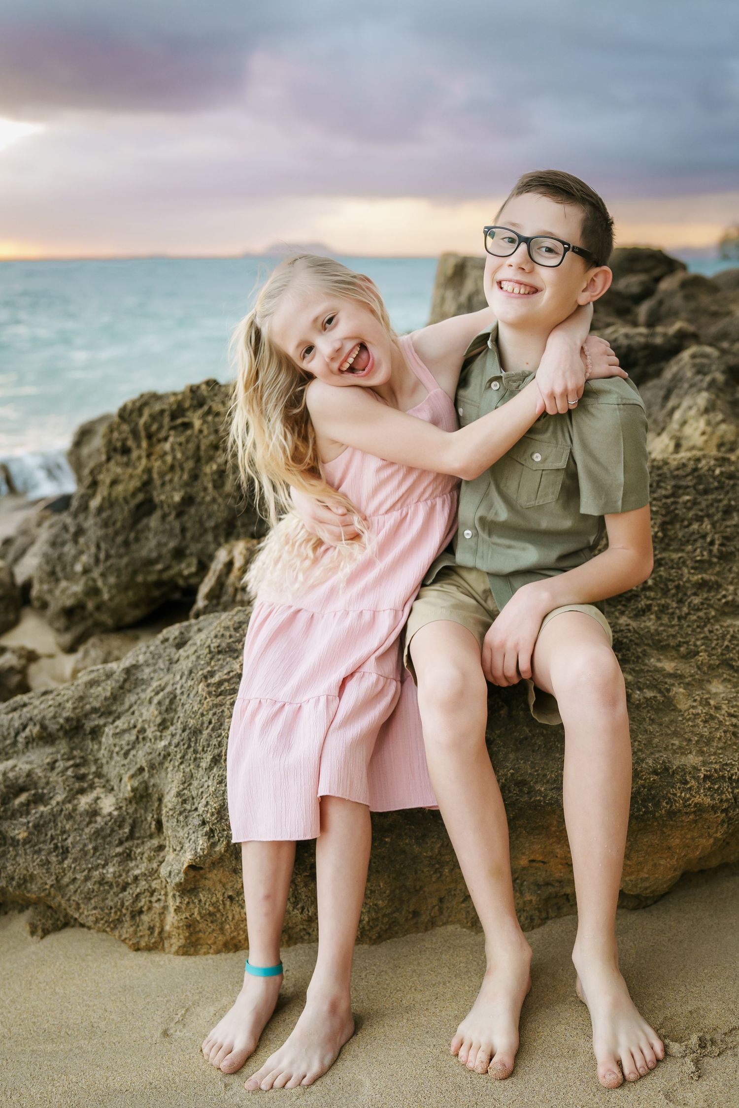 Siblings laughing and hugging on beach rocks at sunrise. Girl in a pink dress and boy in a khaki suit with glasses, with the ocean behind.