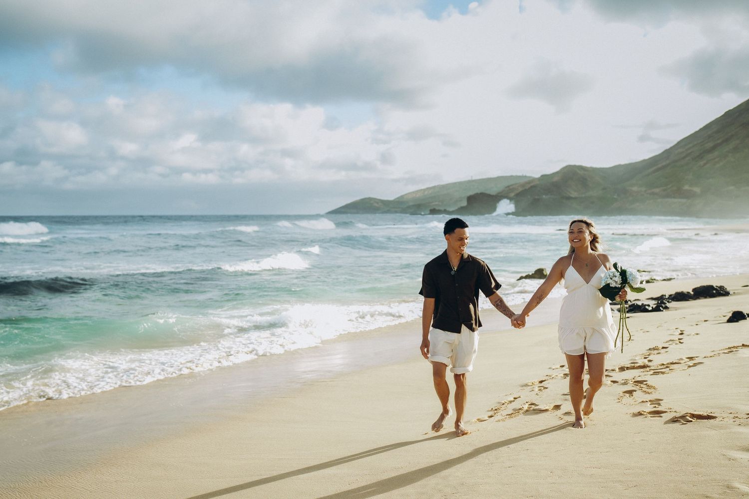 Couple holding hands walking on a sandy beach with turquoise waves and green hills in the background. Anna Tsenta family photographer