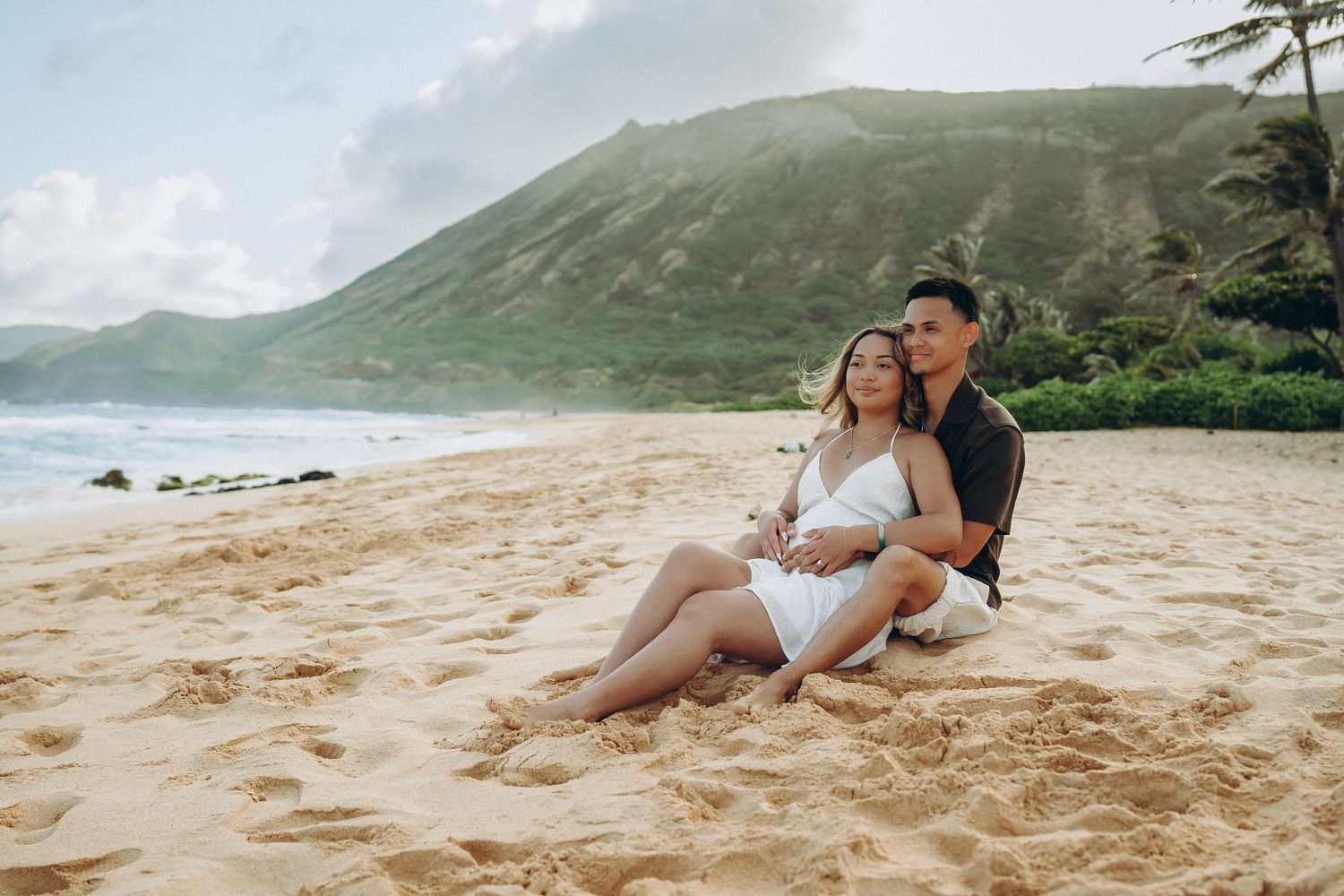 Couple sitting together on sandy beach with lush green mountains and ocean in background during golden hour at Sandy beach Oahu Hawaii
