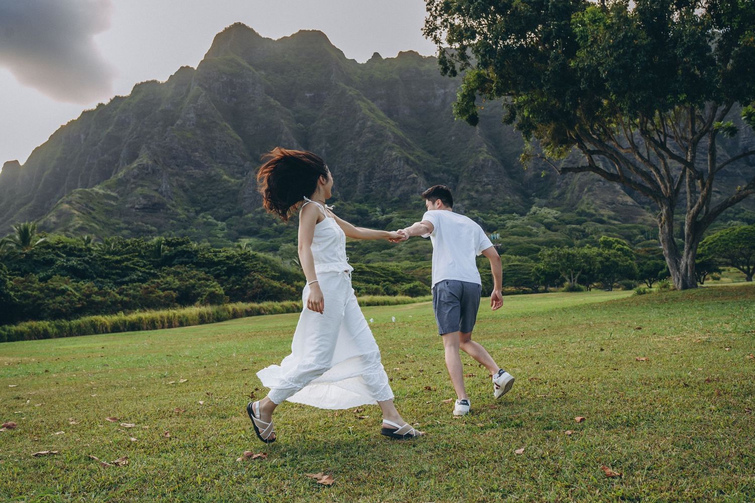 Two people in white clothing run playfully across a grassy field with dramatic mountain peaks at Kualoa Ranch Regional park