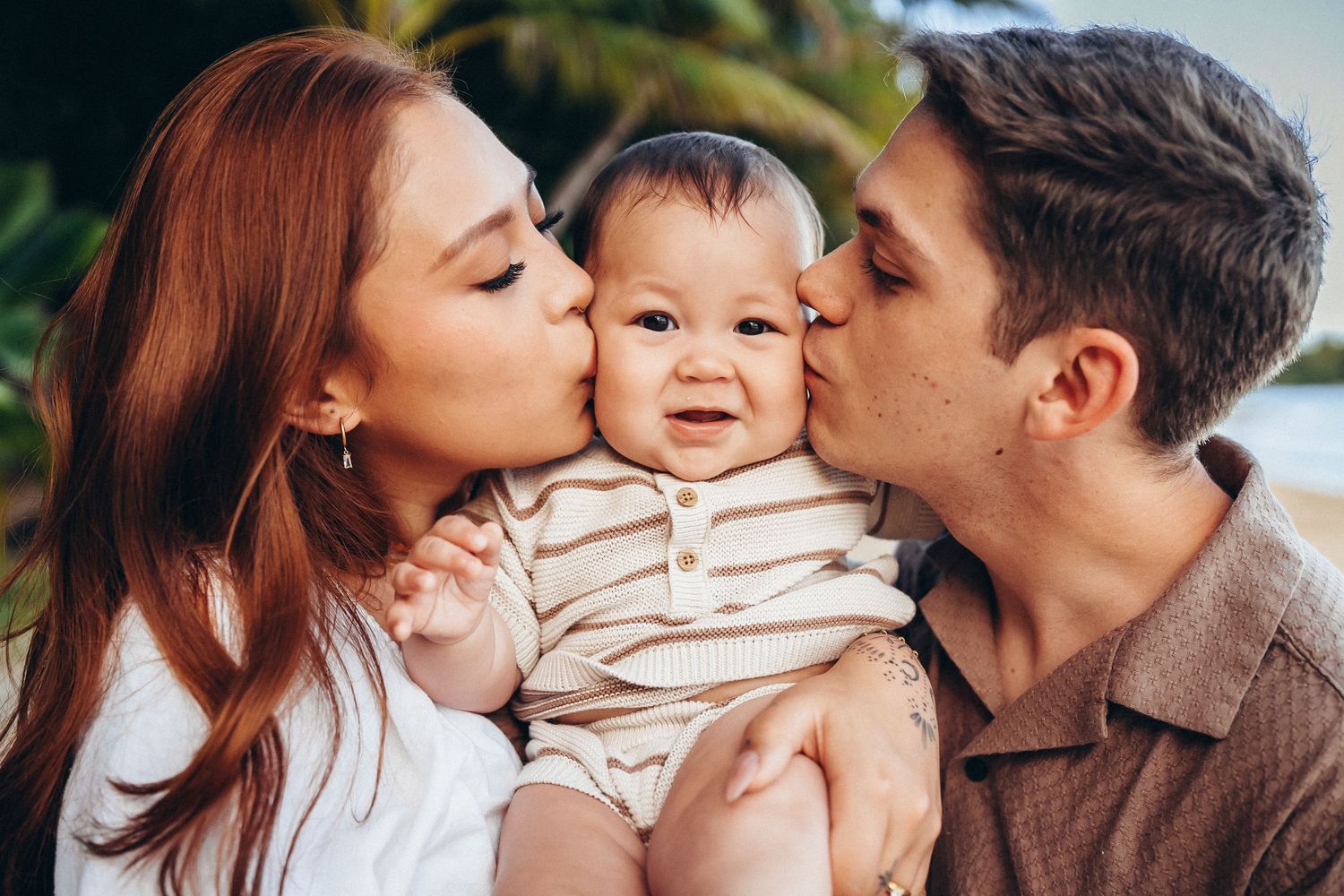 Family photoshoot at Kualoa Ranch, Oahu. Parents kissing their baby's cheeks. Loving family moments at Secret Beach photo session.