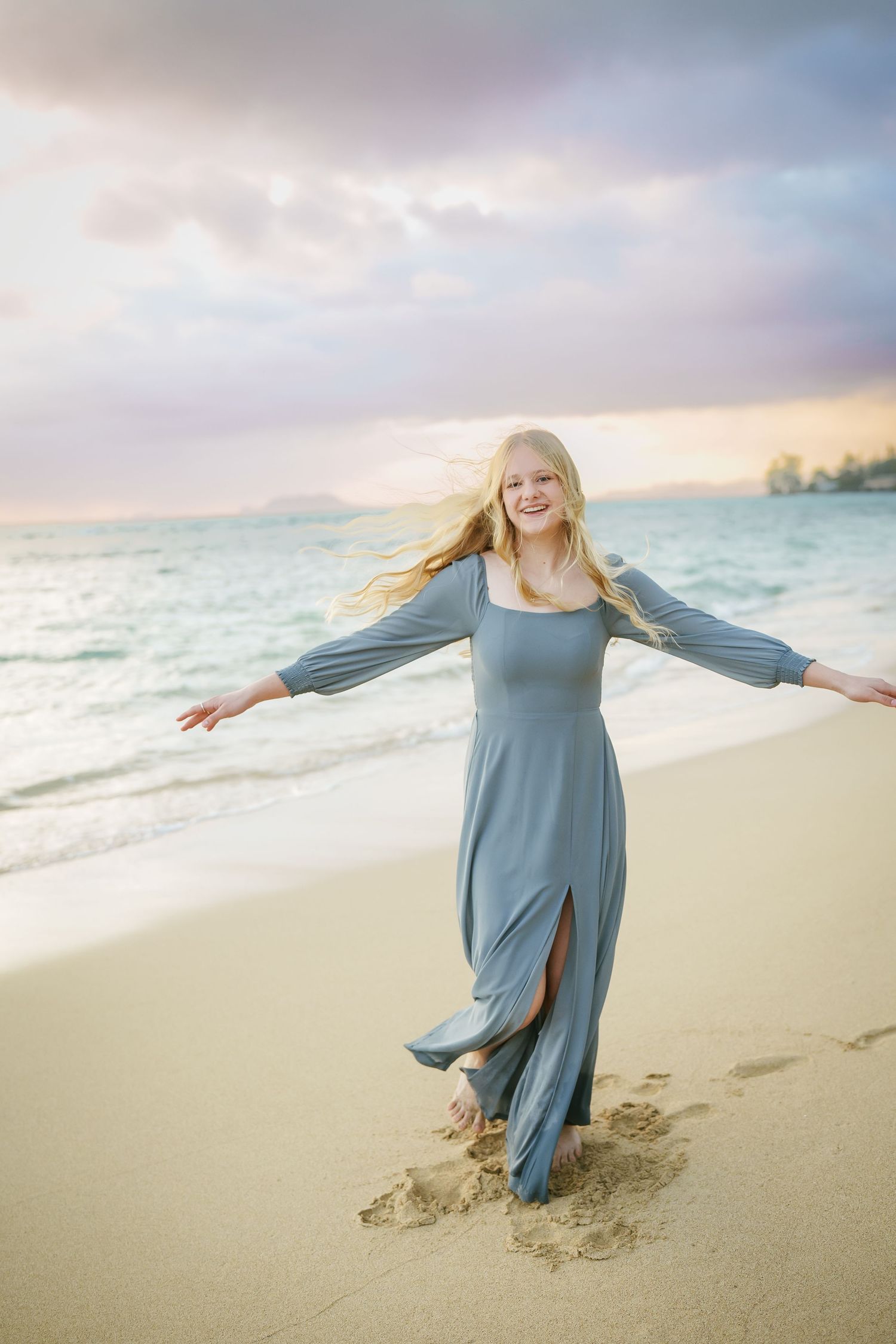 A girl in a long blue dress twirling on the white sand at sunrise. A graceful, cinematic moment at Ka’a’awa Beach, Oahu, with the ocean behi