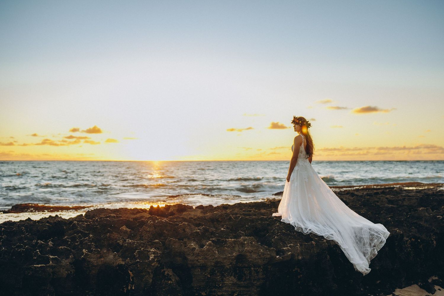 Bride in a white wedding dress standing on the lava rock shore at sunset, Four Seasons Resort O'ahu at Ko Olina.