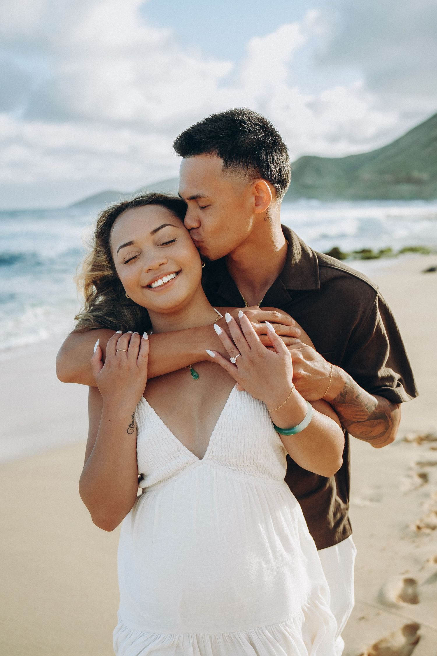 A couple embraces on a sandy beach, the man kissing the smiling woman's cheek with ocean waves behind them at Sandy beach Oahu Hawaii