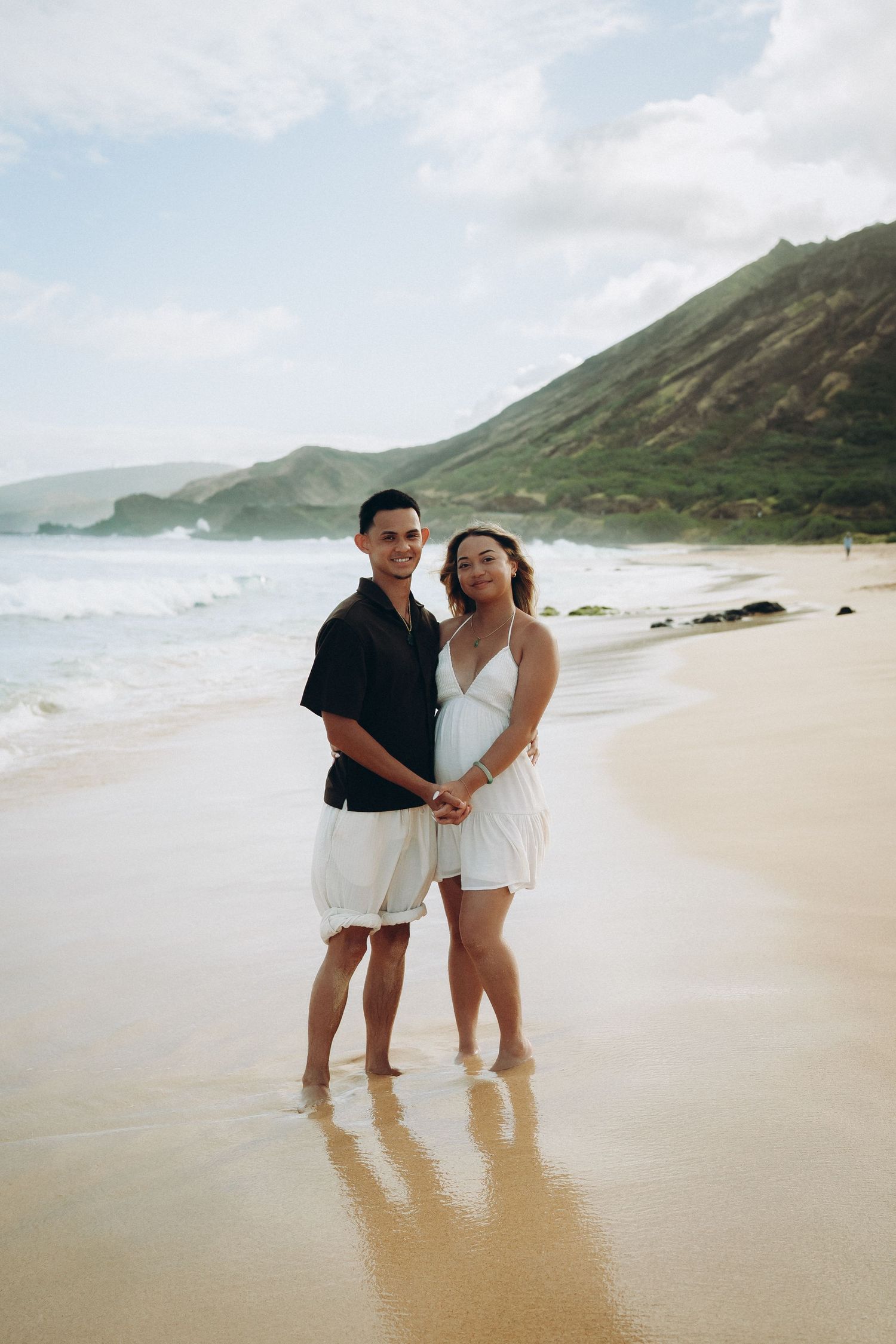 Couple holding hands on a sandy beach with ocean waves and green mountains in the background on a cloudy day at Sandy beach Oahu Hawaii
