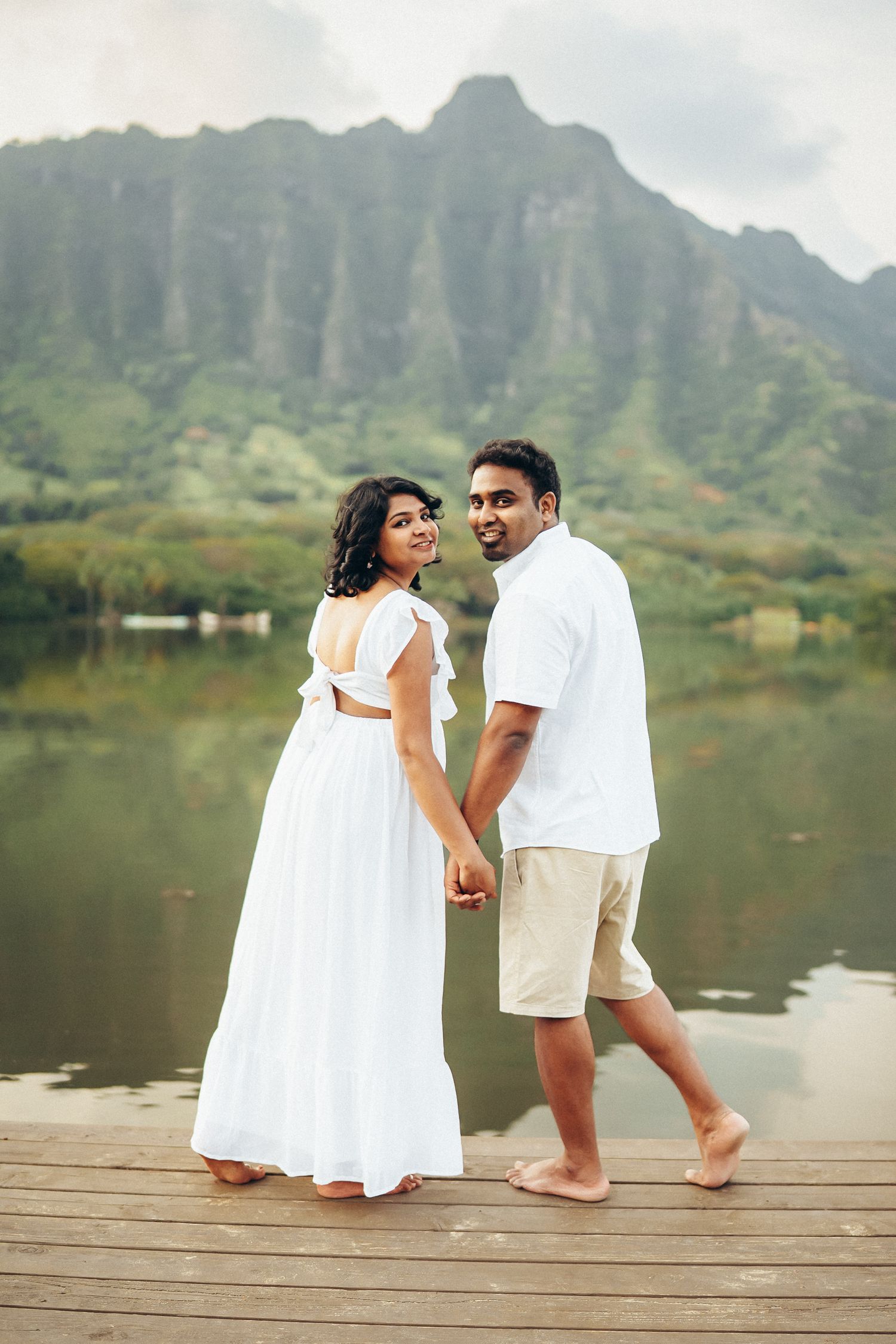 Family photoshoot at Kualoa Ranch, Oahu. A couple holding hands on a pier at Secret Beach. Stunning mountain backdrop and calm water.
