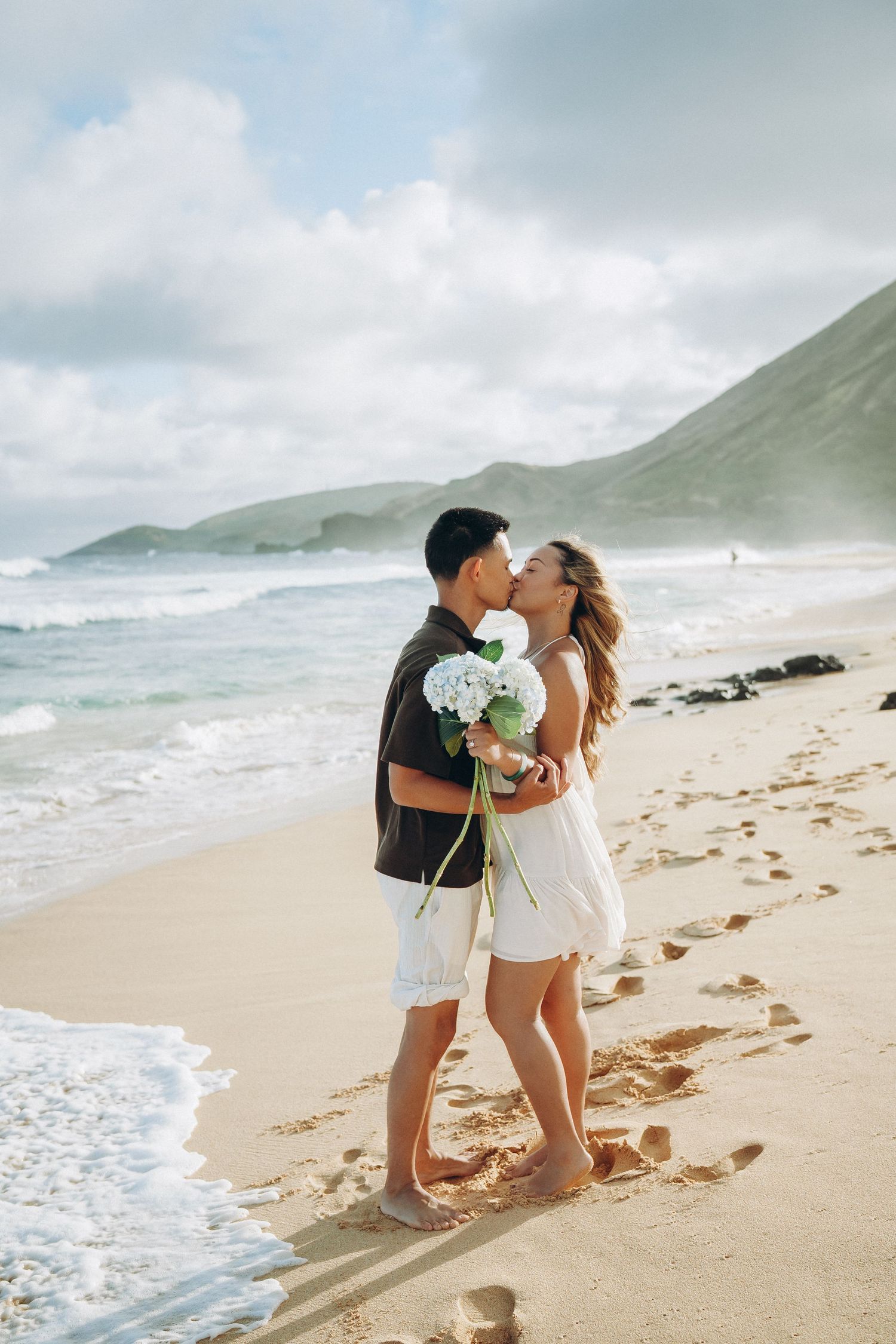 Couple kissing on a sandy beach holding white flowers with ocean waves and green mountains in the background at Sandy beach Oahu Hawaii