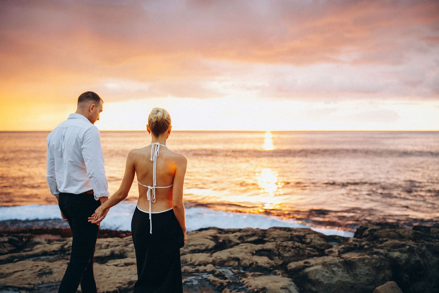 Ko Olina engagement. Romantic couple silhouetted against vibrant orange sunset sky on rocky Hawaiian coastline during golden hour.