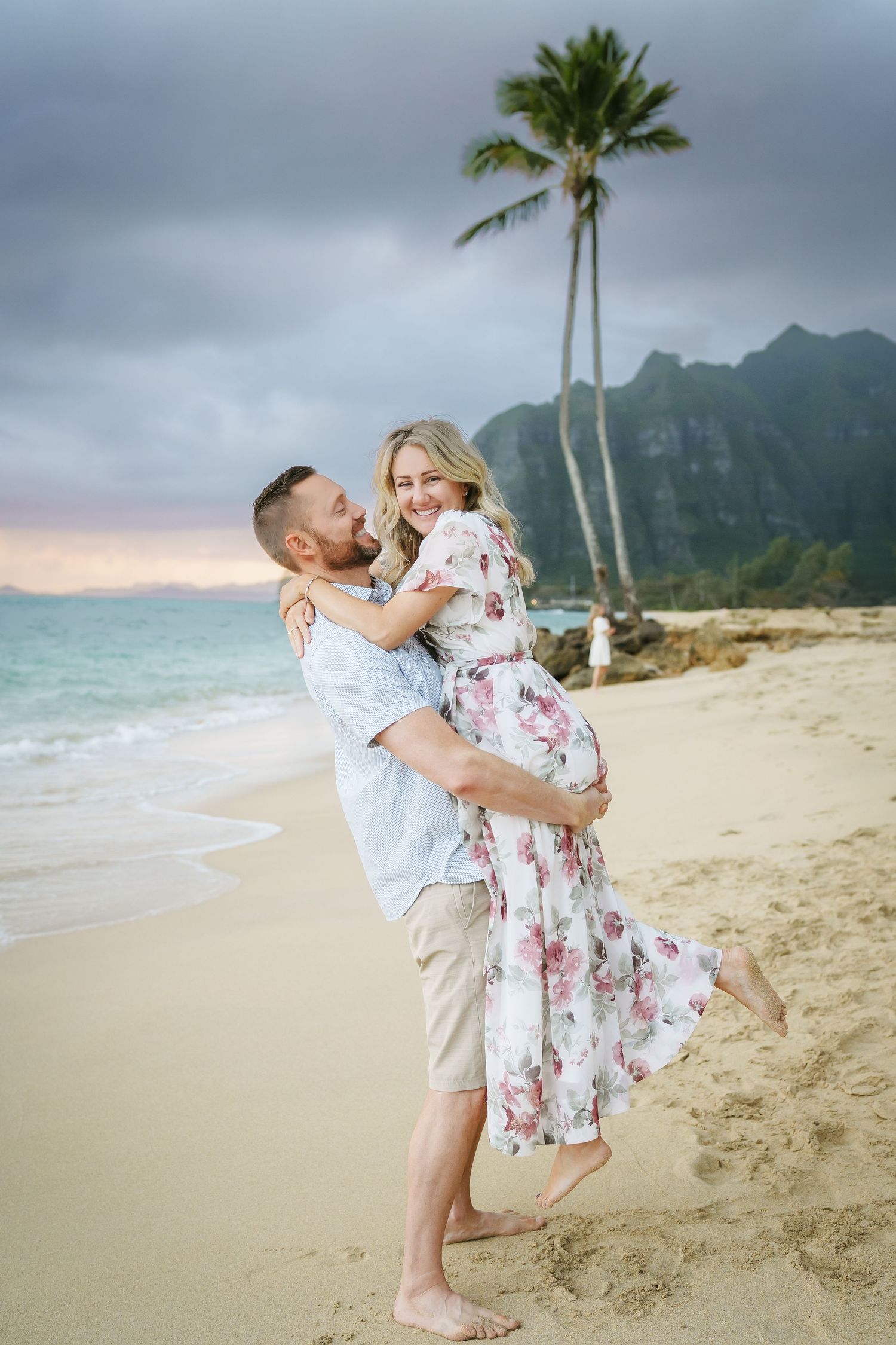 Parents embracing as the father lifts and twirls the mother at Ka’a’awa Beach, Oahu. A romantic moment with palm trees, mountains, and ocean