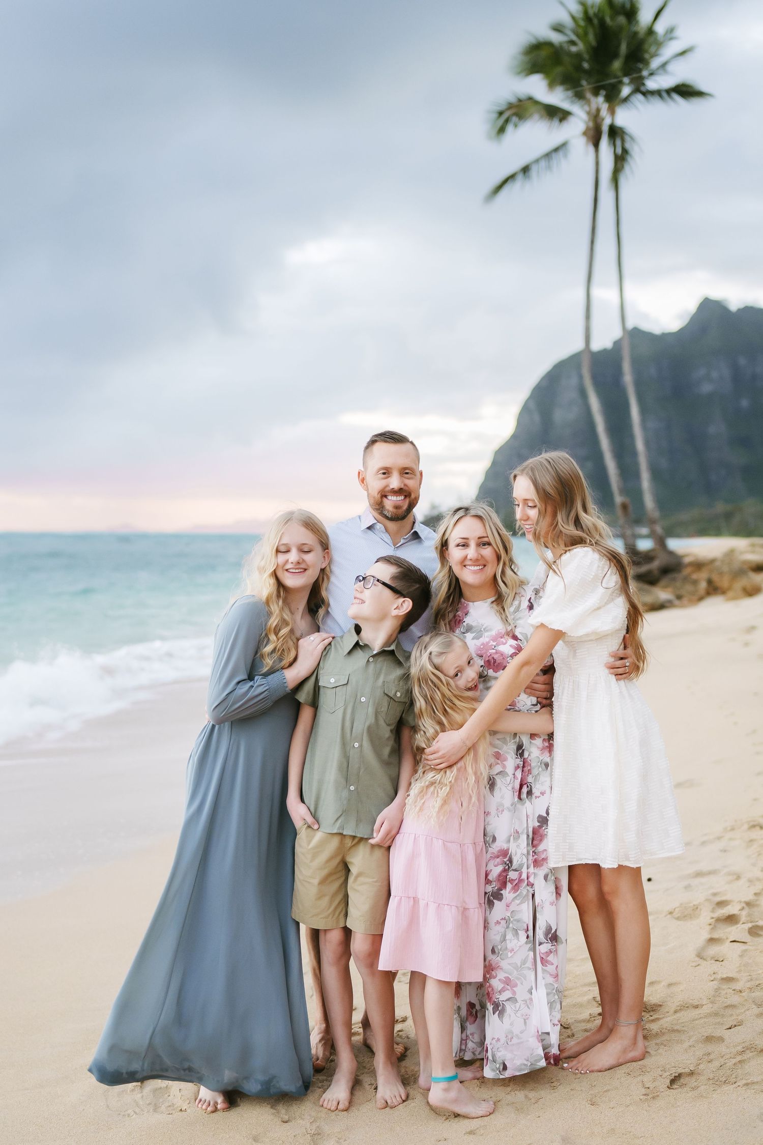 A tender moment with a family of six smiling and looking at each other at Ka’a’awa Beach, Oahu. Scenic mountains, palm trees, and ocean in t