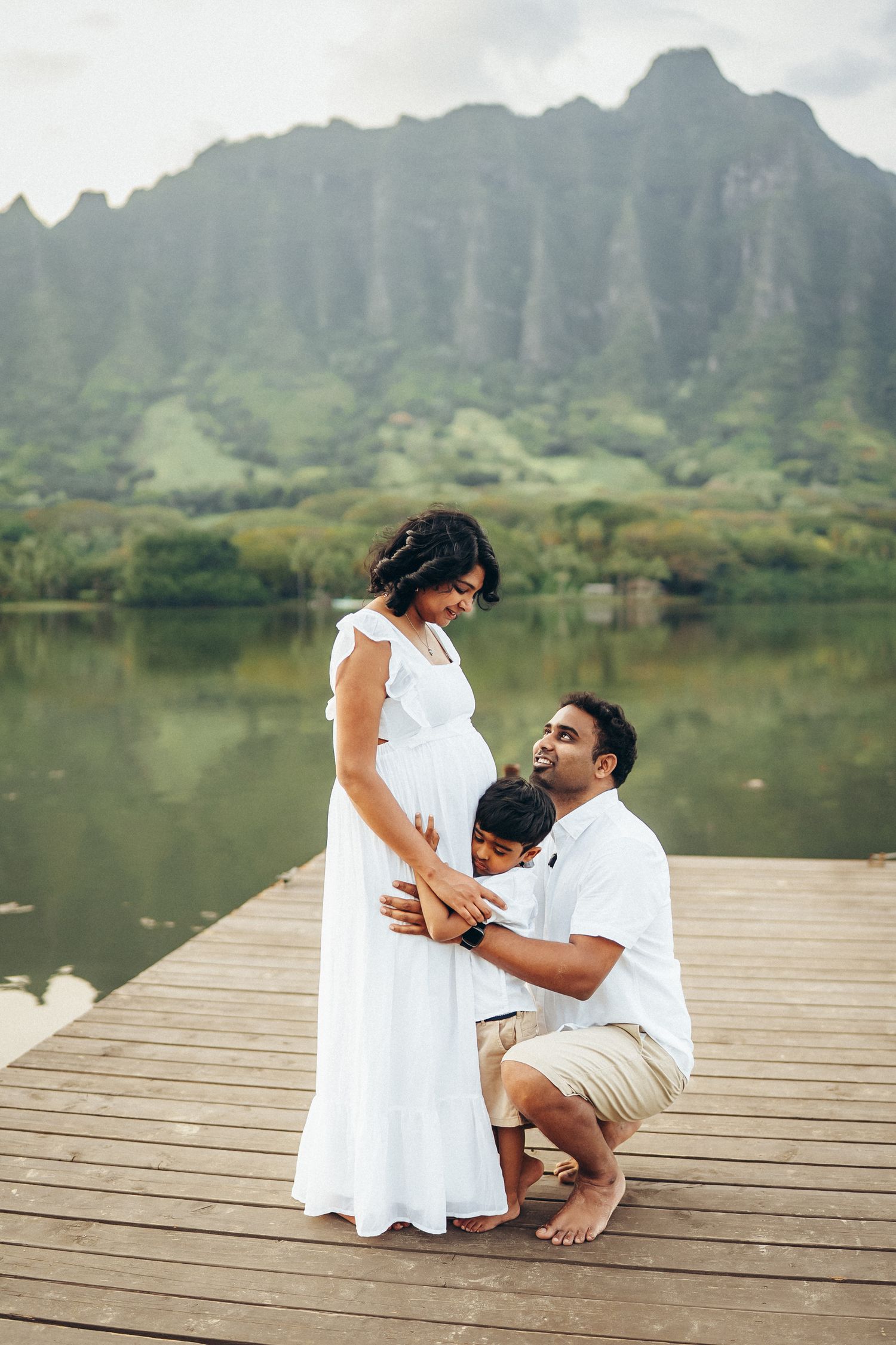 Oahu family photoshoot at Secret Beach. Husband kissing pregnant wife's belly on a pier with Kualoa Ranch mountains in the background.