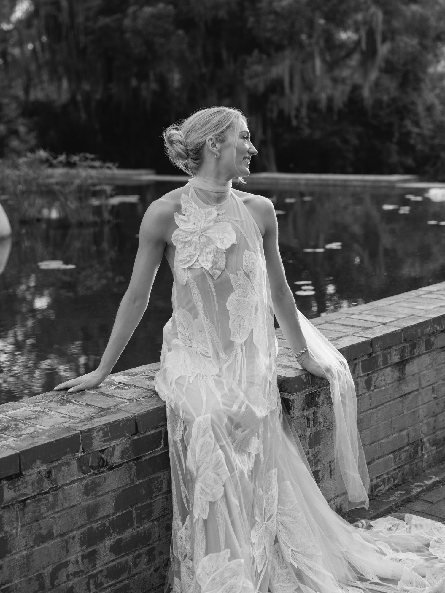 Bride in floral appliqué halter wedding gown poses by brick wall near pond in elegant black and white portrait.