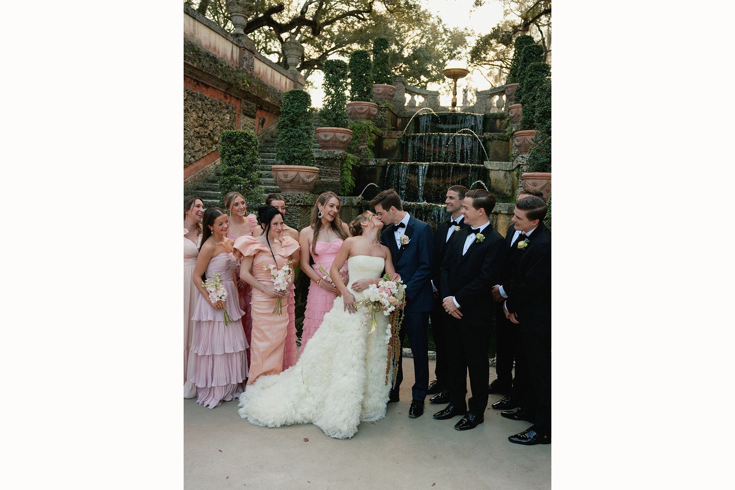 Wedding party members in pink dresses and dark suits pose together outdoors for formal group photos.