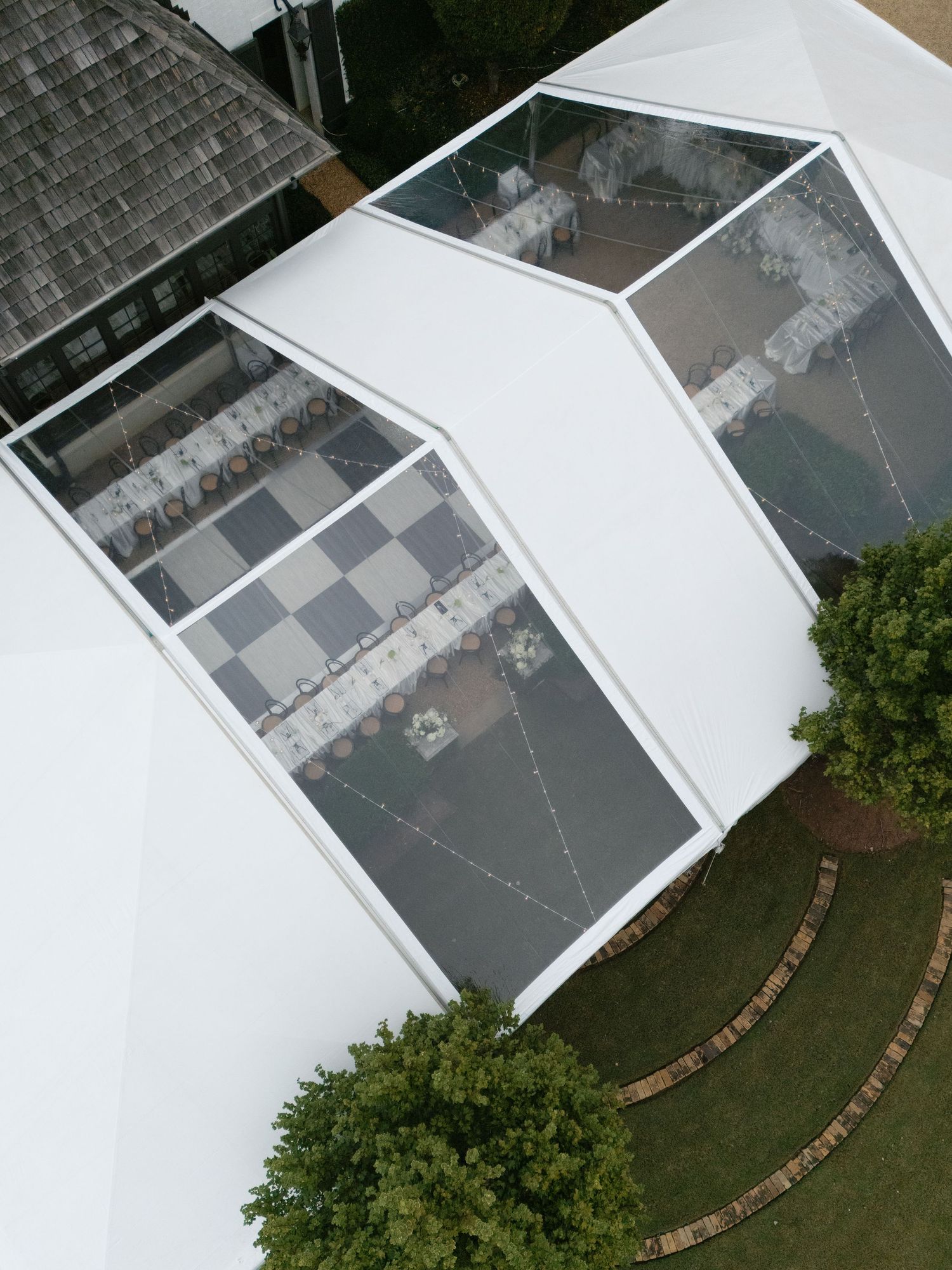 Aerial view of a white wedding tent with clear panels showing tables set up in a checkerboard pattern on grass.