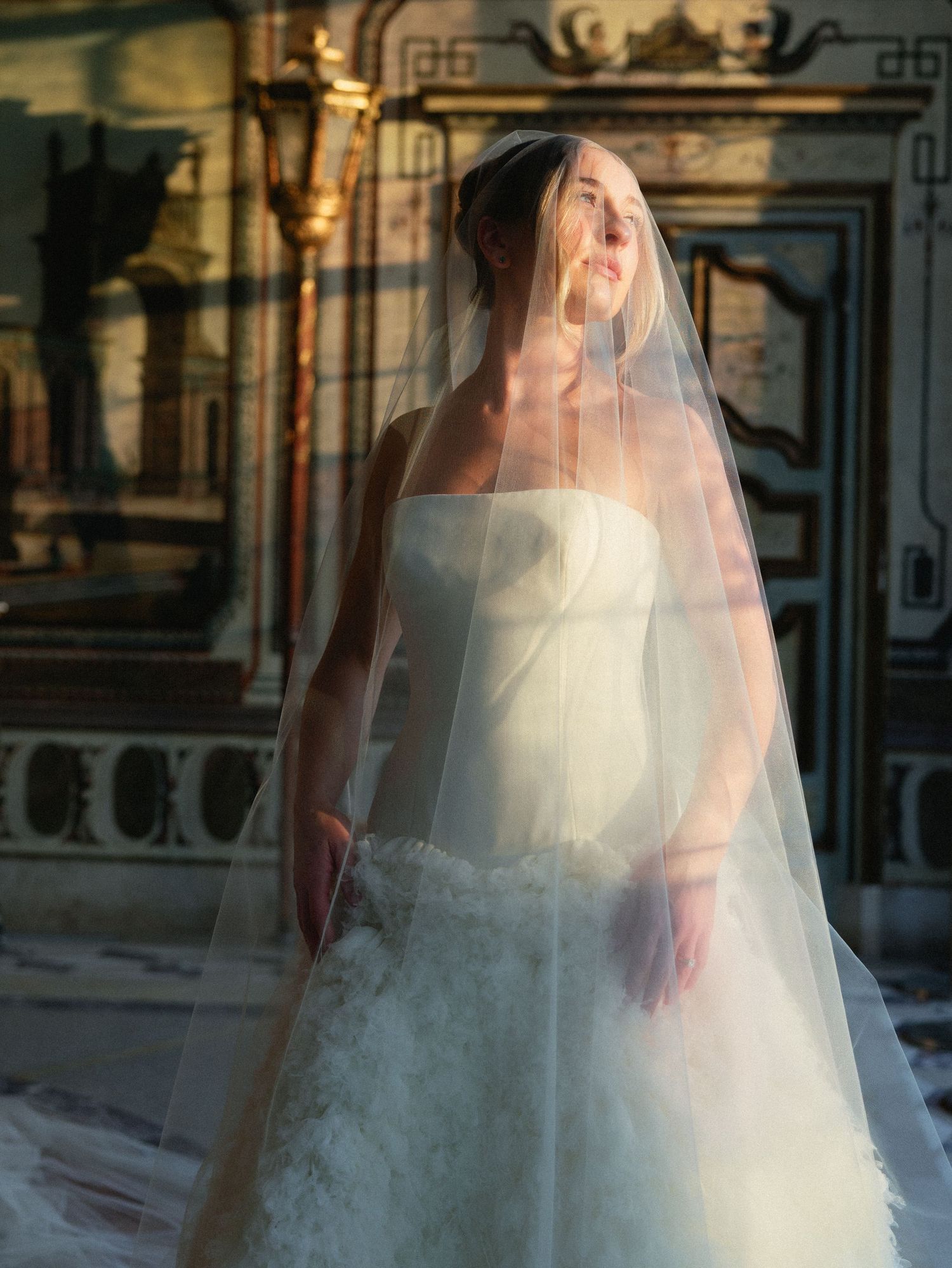Bride in strapless white gown and flowing veil stands in golden light inside an ornate classical venue.