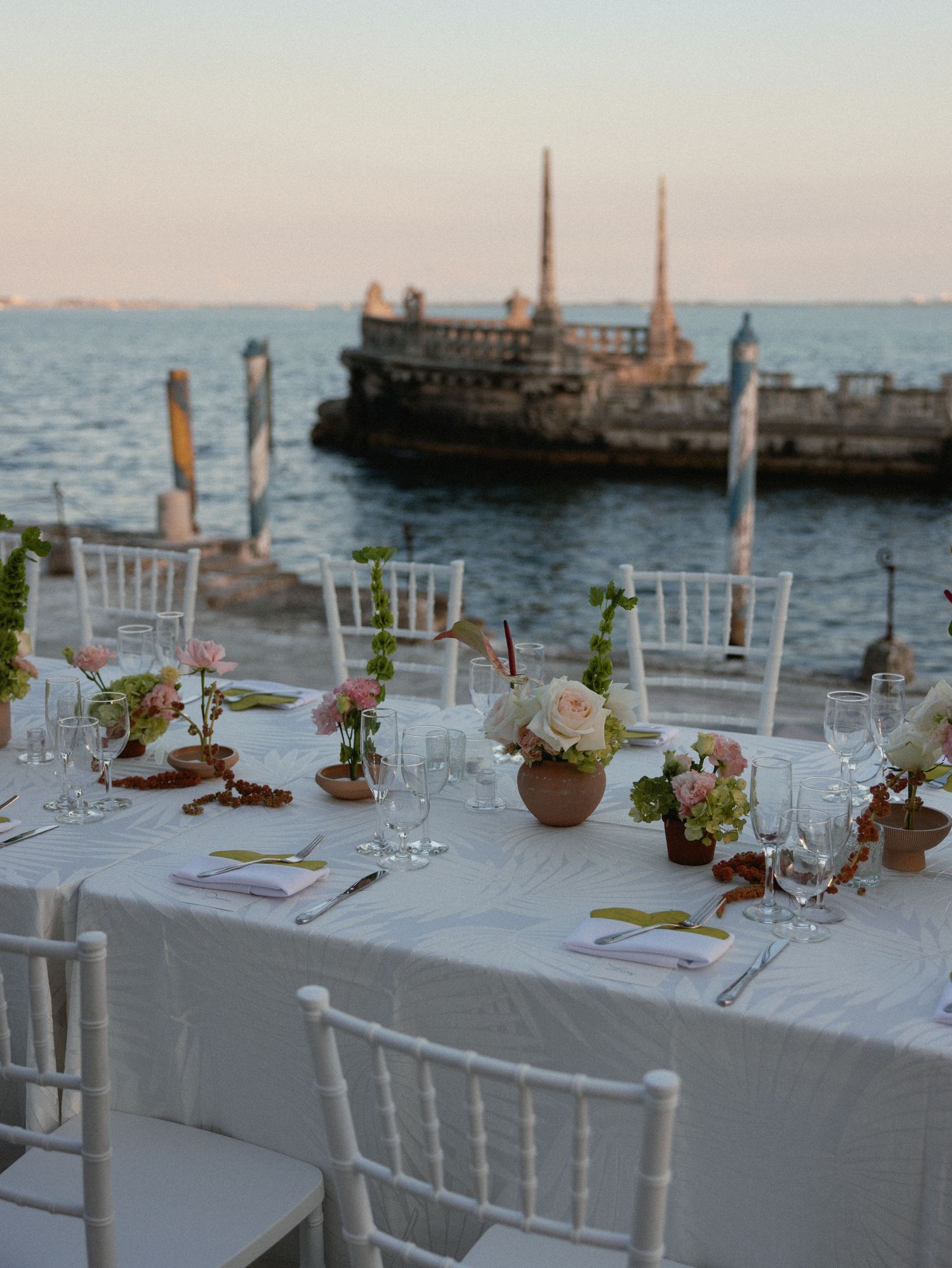 Elegant seaside dinner table setting with white chairs and floral centerpieces overlooking a docked boat at sunset.