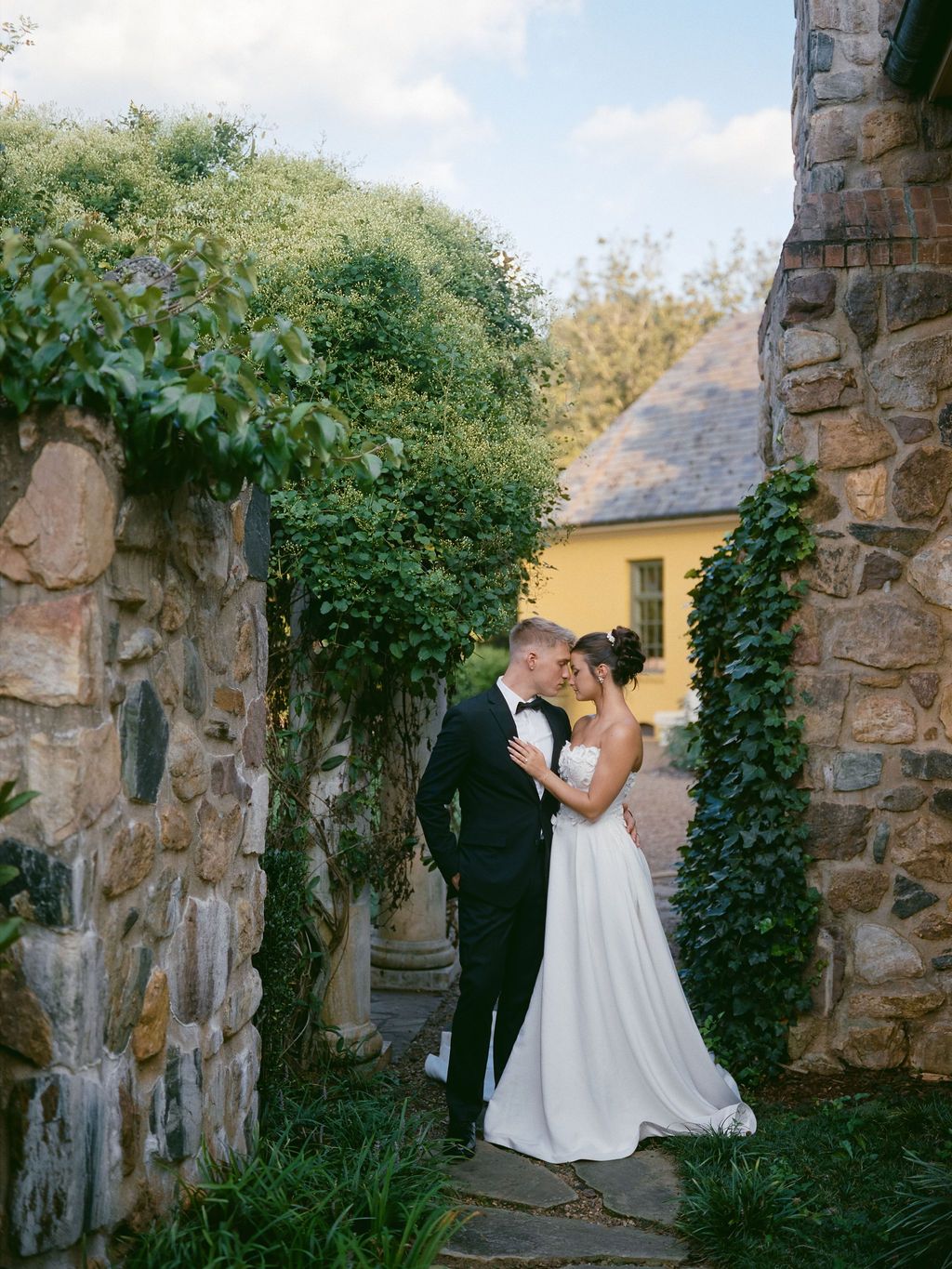 A romantic wedding moment captured between stone walls covered in ivy and a yellow building in the background.
