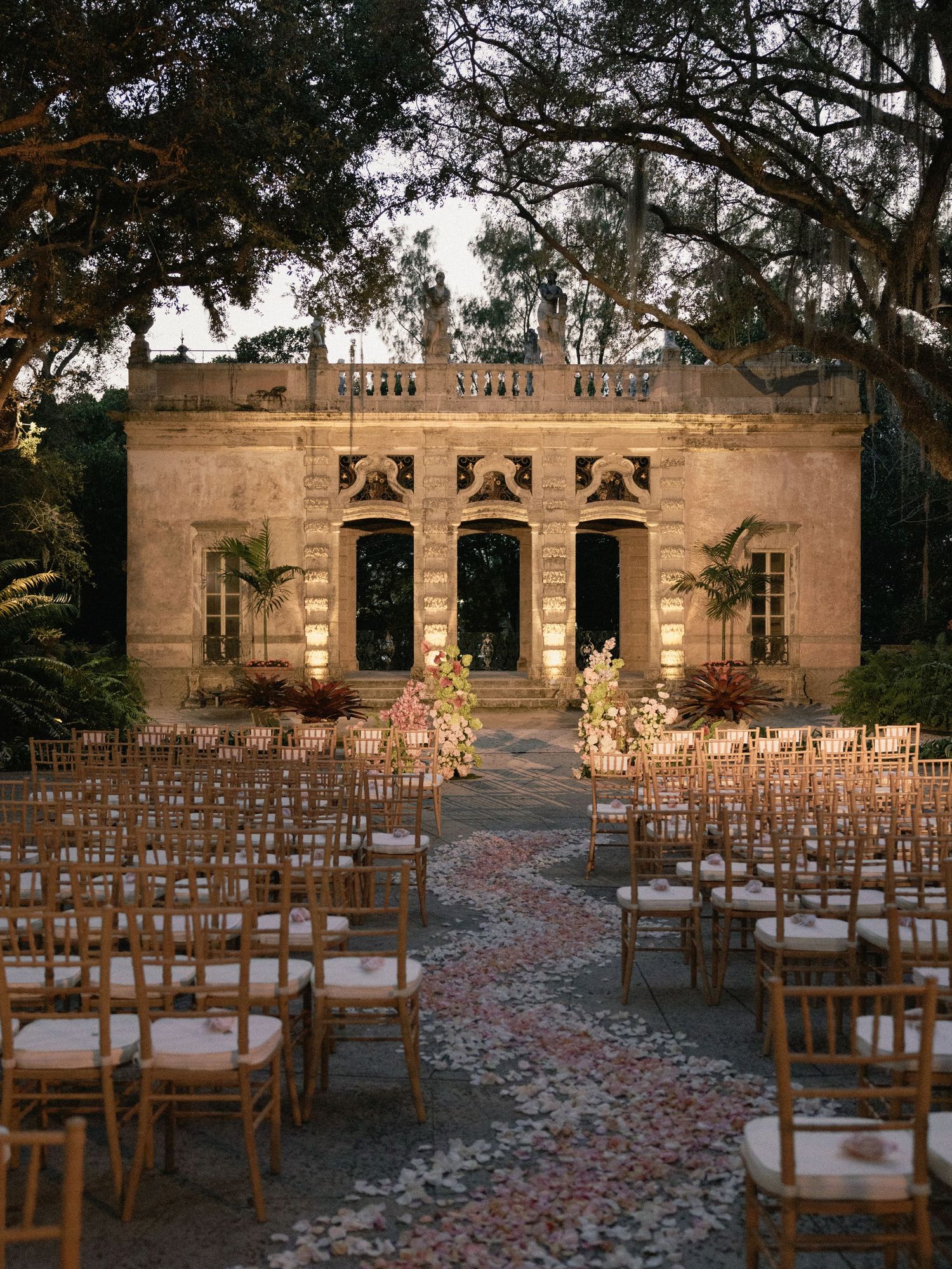 Outdoor wedding ceremony setup with wooden chairs arranged in rows along a rose petal aisle leading to a stone building.