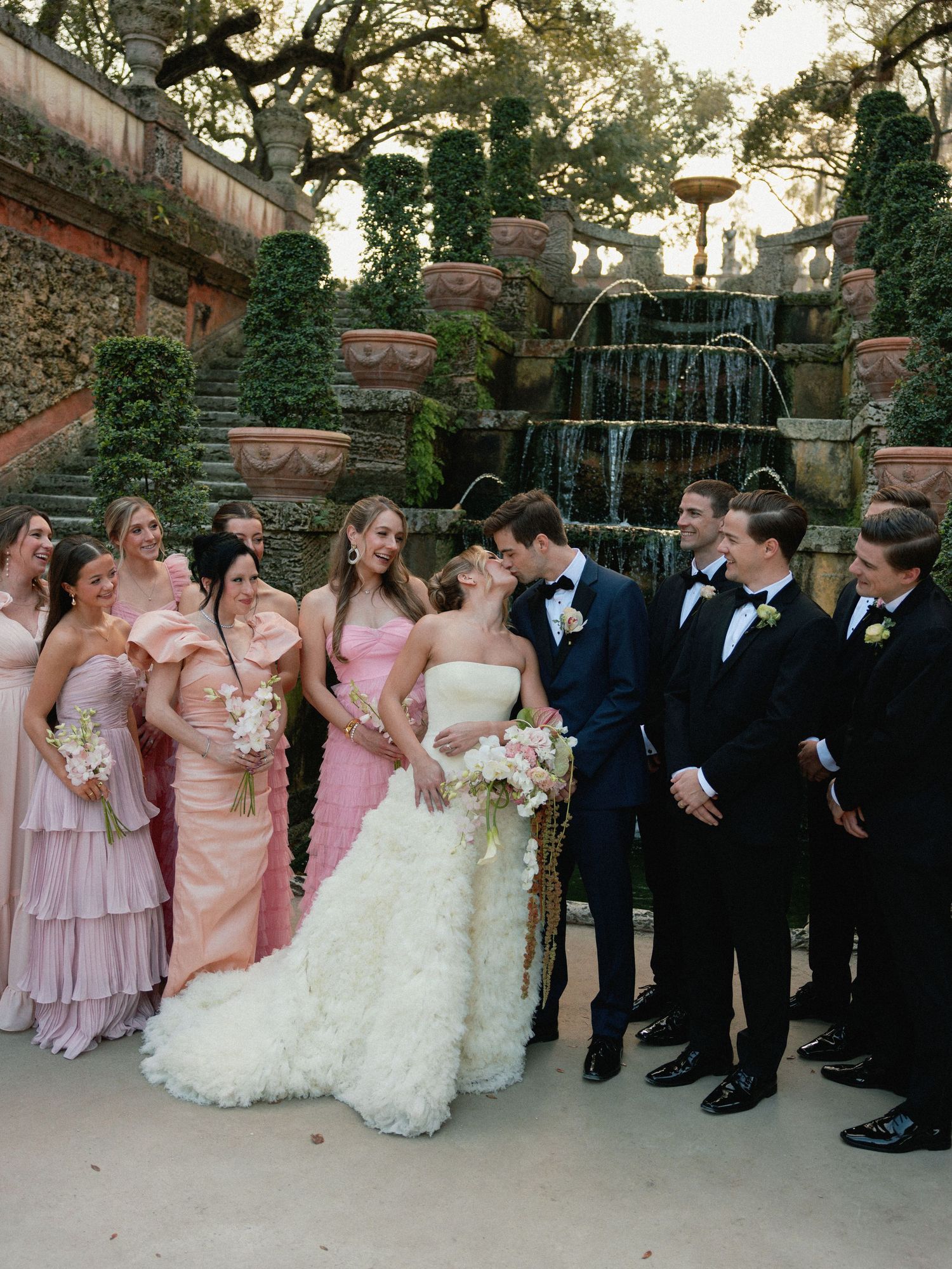 Wedding party in pink dresses and black tuxedos poses together in a garden setting at sunset.