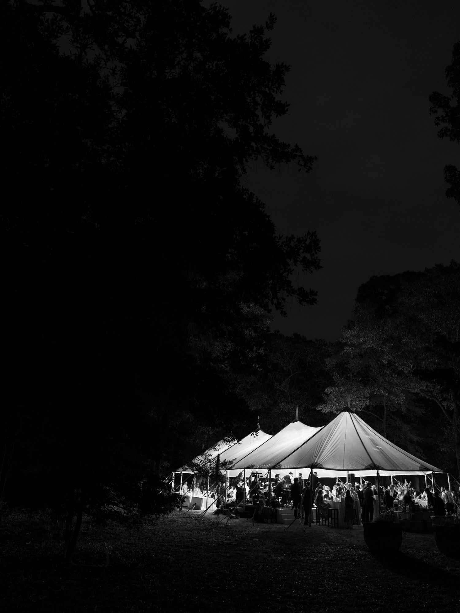Glowing wedding reception tent illuminated at night surrounded by dark trees with guests gathered outside.